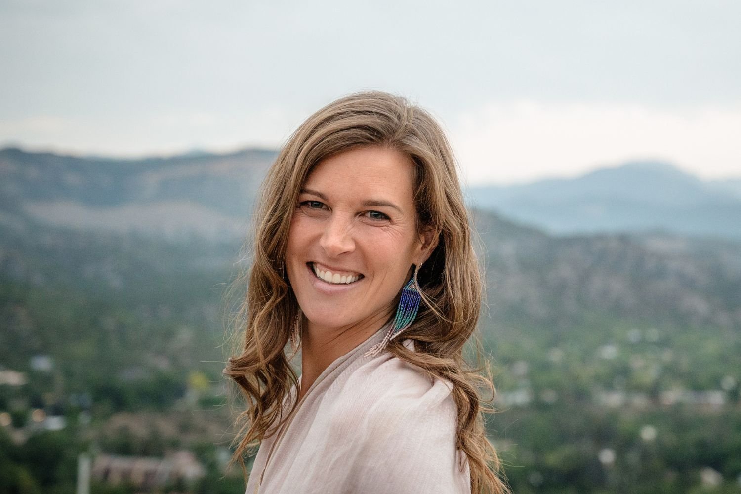 A woman smiling outdoors with mountains and a valley in the background, wearing a beige top and colorful feather earrings.