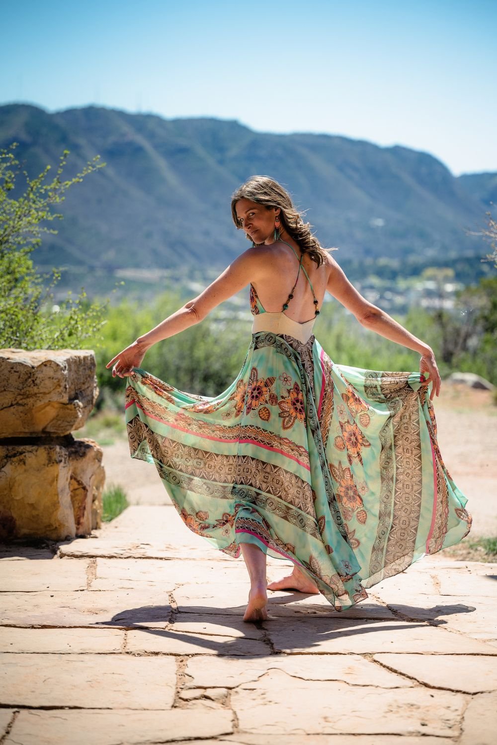 A woman in a flowing, colorful dress poses outdoors with mountains and trees in the background.