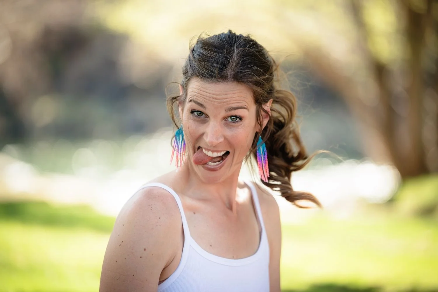 A woman with curly brown hair, wearing colorful, feathered earrings and a white tank top, sticking out her tongue playfully outdoors in a park with a blurred background of trees and sunlight.