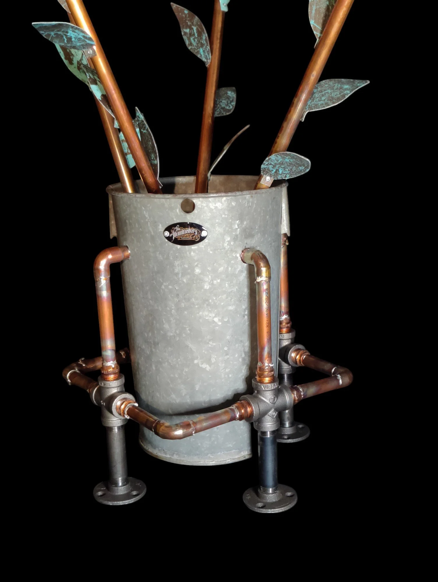 A plant in a metallic container surrounded by copper pipes and fittings, with black background.