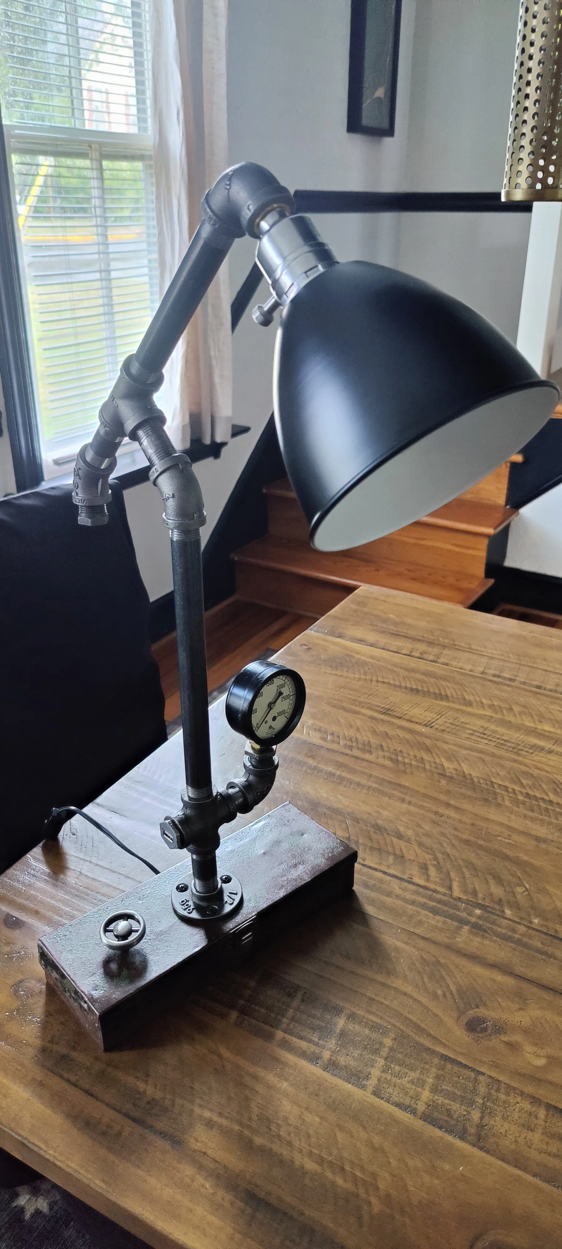 A vintage industrial-style table lamp with a black metal shade, mounted on a wooden table. The lamp features a black pipe structure with a pressure gauge at the base, and is positioned near a window with white blinds, in a room with wooden stairs and modern decor.