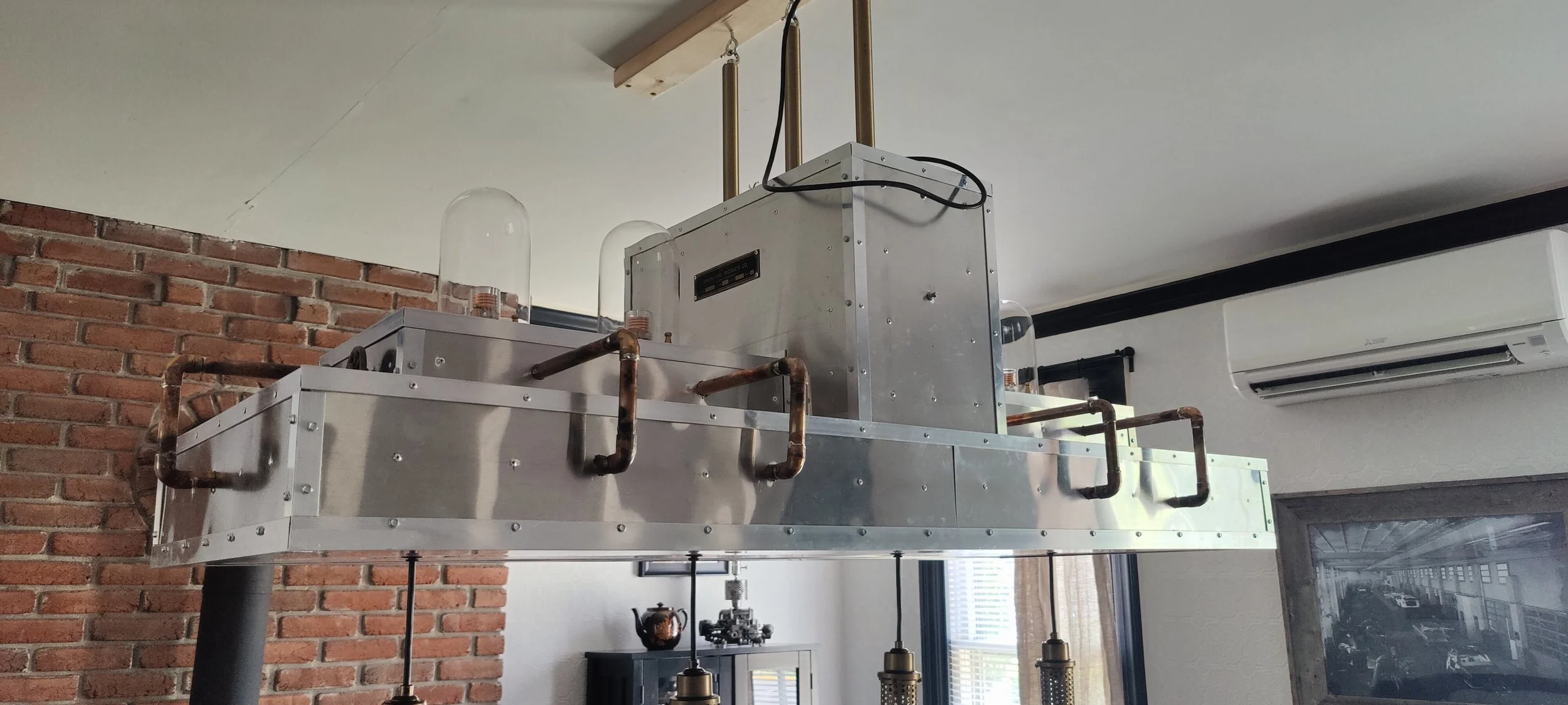 A metallic industrial-style chandelier with visible copper pipes, hanging in a room with a brick wall, air conditioning unit, and framed black and white photograph.