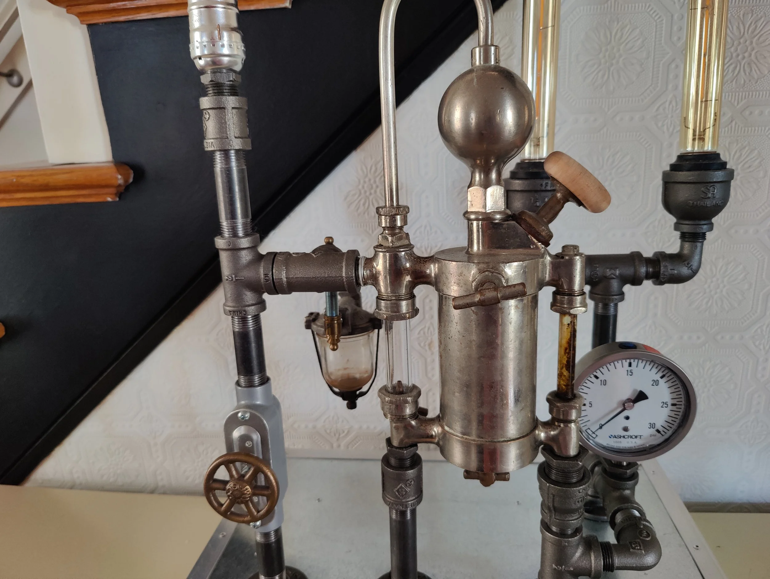 A laboratory setup with pipes, valves, and a pressure gauge, featuring a metal sphere on top and a glass container with fluid.