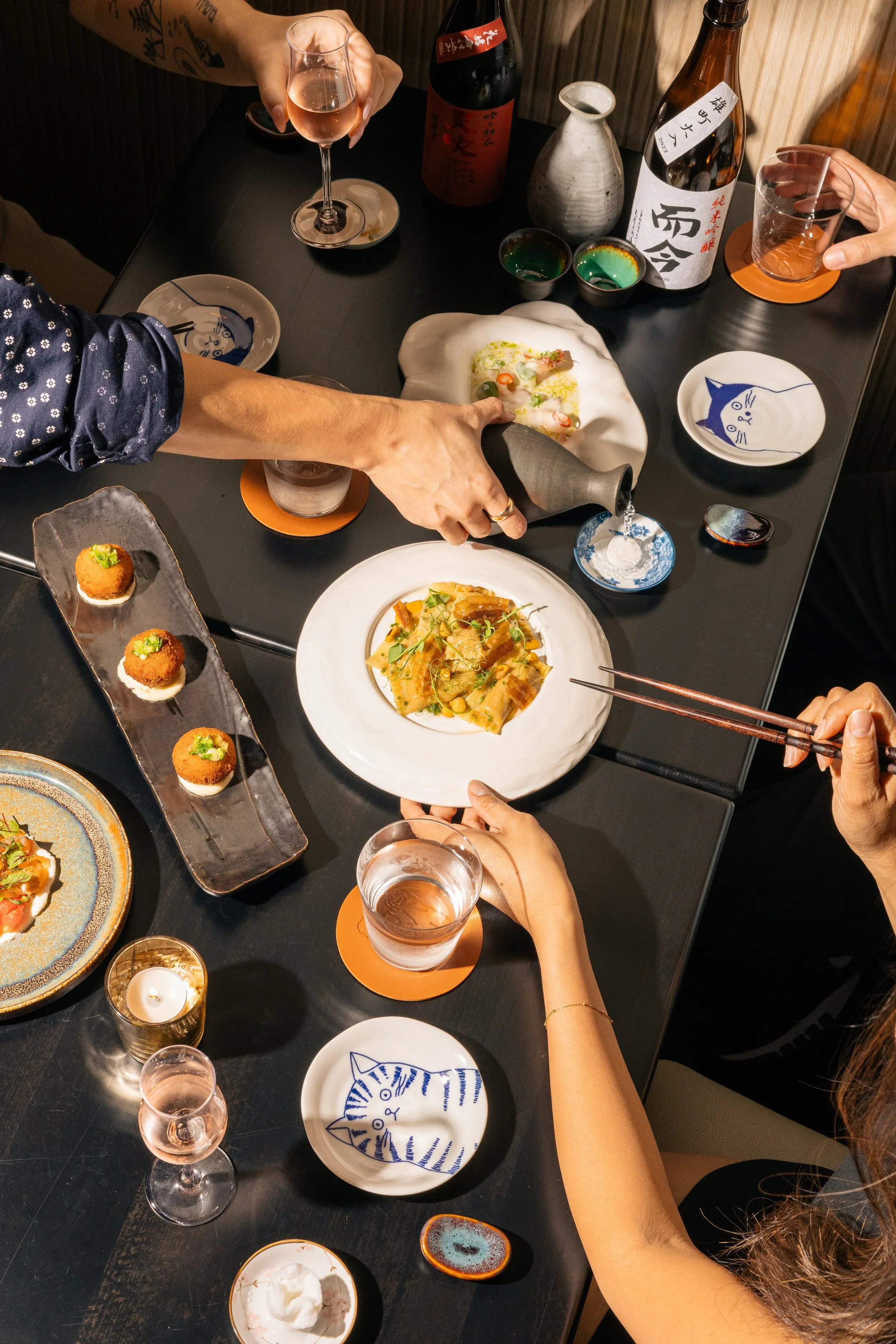 Overhead view of diners enjoying a Japanese meal with assorted dishes, chopsticks, sake bottles, wine glasses, and decorative cat plates.