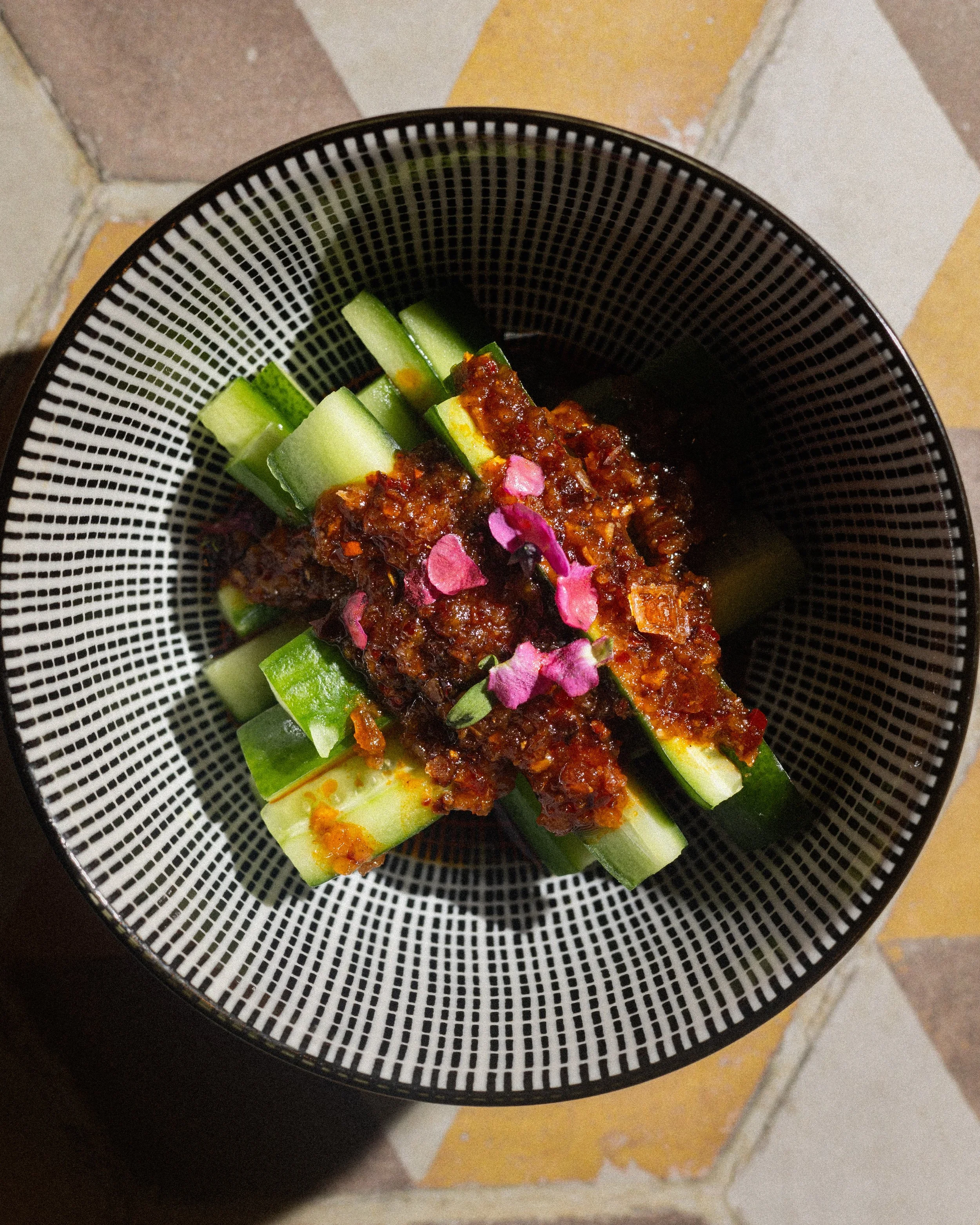 Spicy glazed chicken with cucumber slices and pink flower petals on a black-and-white patterned plate