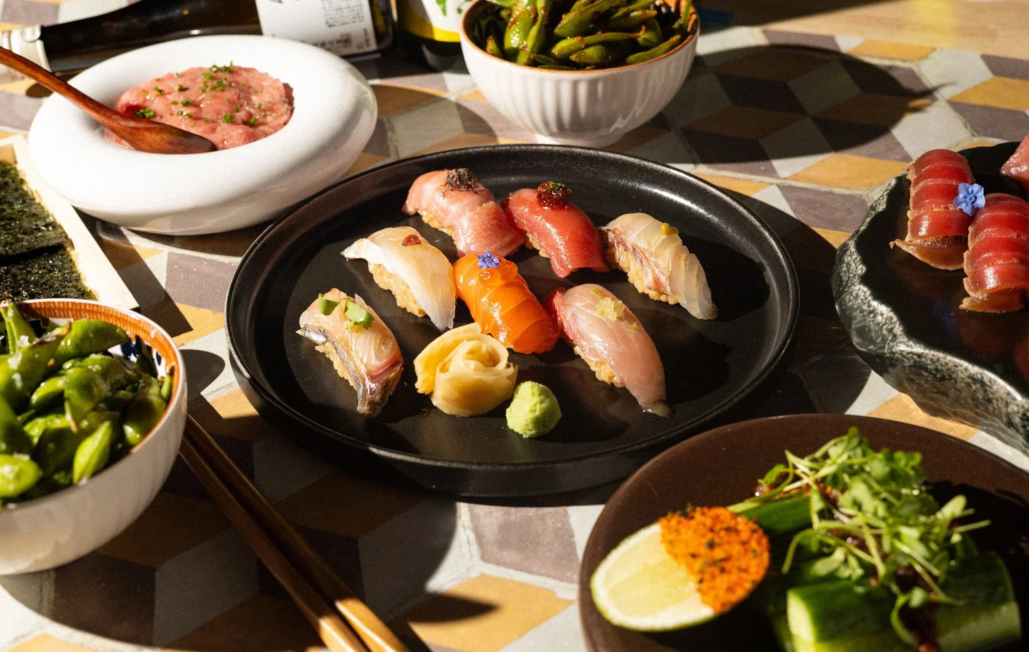 Assorted sushi nigiri on a black plate with edamame, sashimi, wasabi, pickled ginger, and side dishes