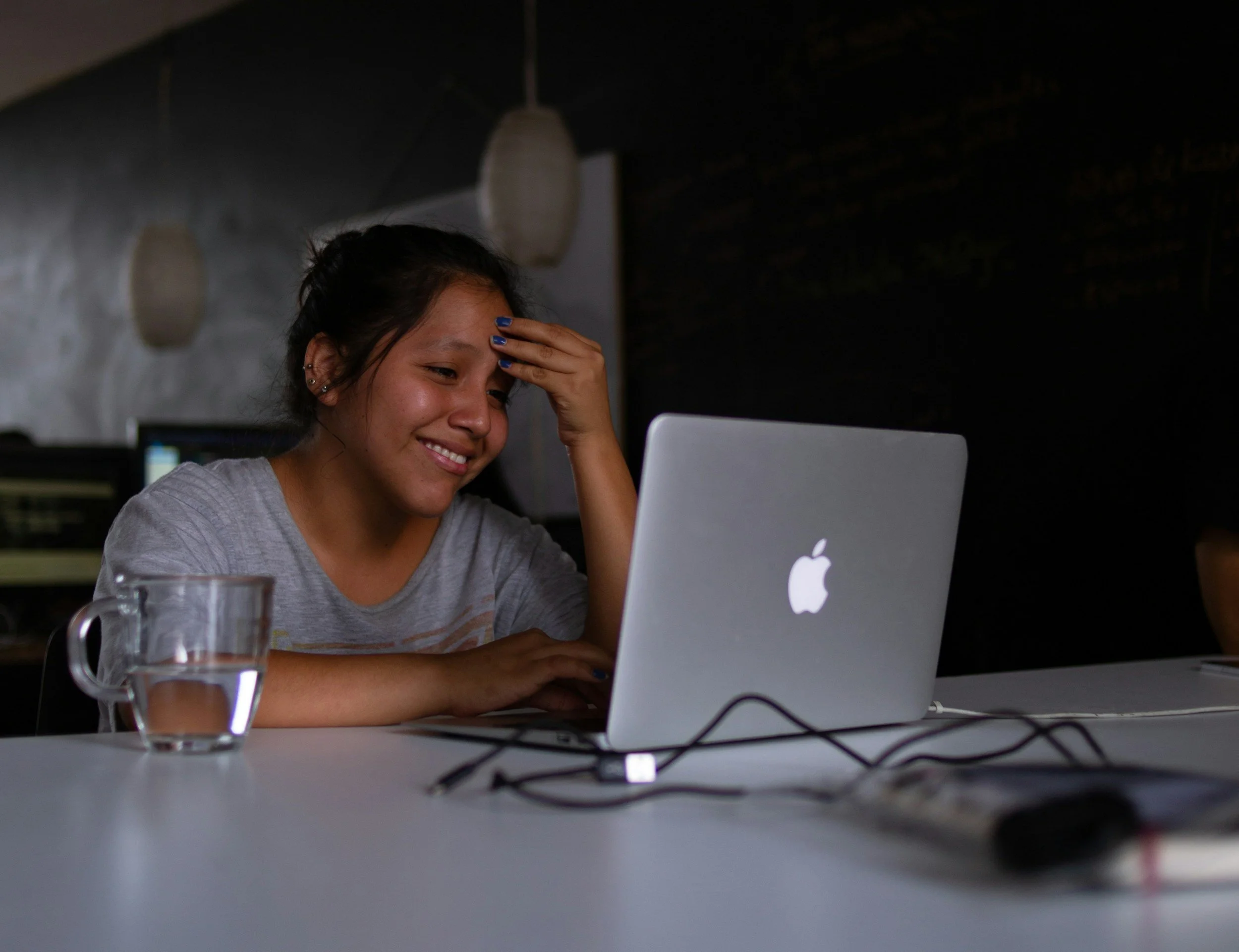 A young woman sitting at a white table, smiling and holding her forehead with her left hand, looking at a silver Apple laptop in front of her. There is a glass cup with water on the table, and the background is dimly lit with pendant lights hanging from the ceiling.