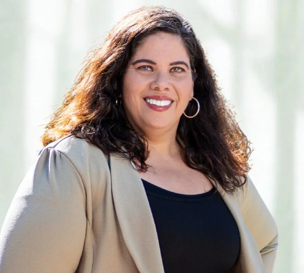 A woman with dark curly hair wearing a beige blazer over a black top, smiling with a neutral background.
