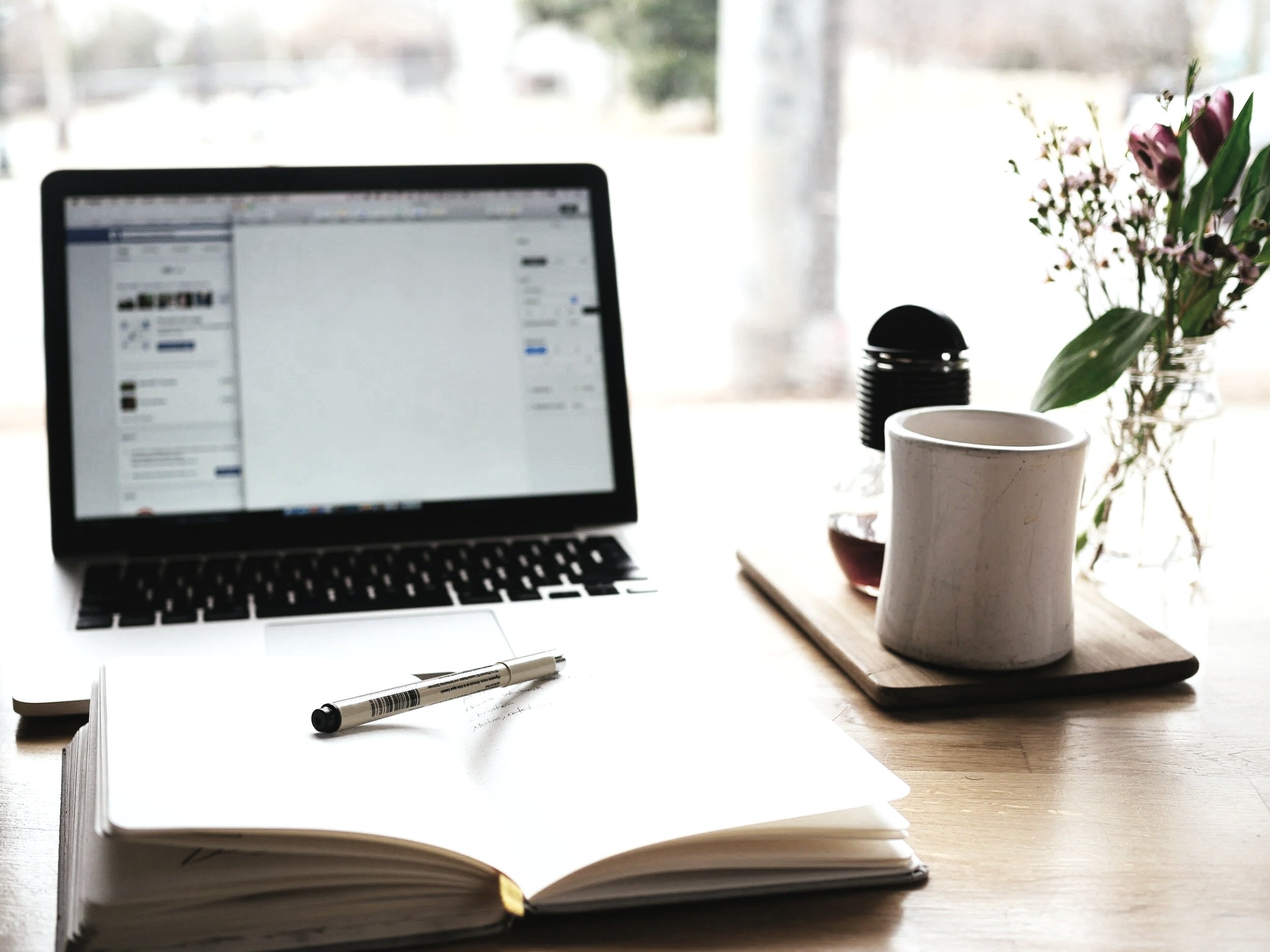 A workspace with an open notebook, a pen, a laptop, a cup, a glass jar with flowers, and a small plate, set on a wooden table near a window.