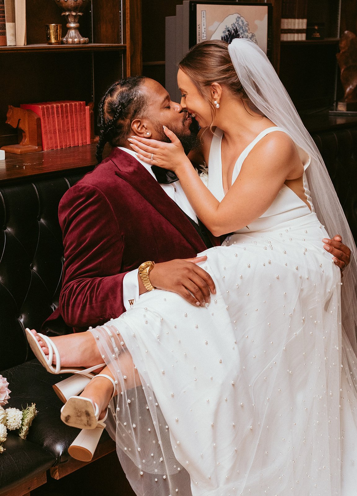 A bride sits on lap of her groom in a lounge