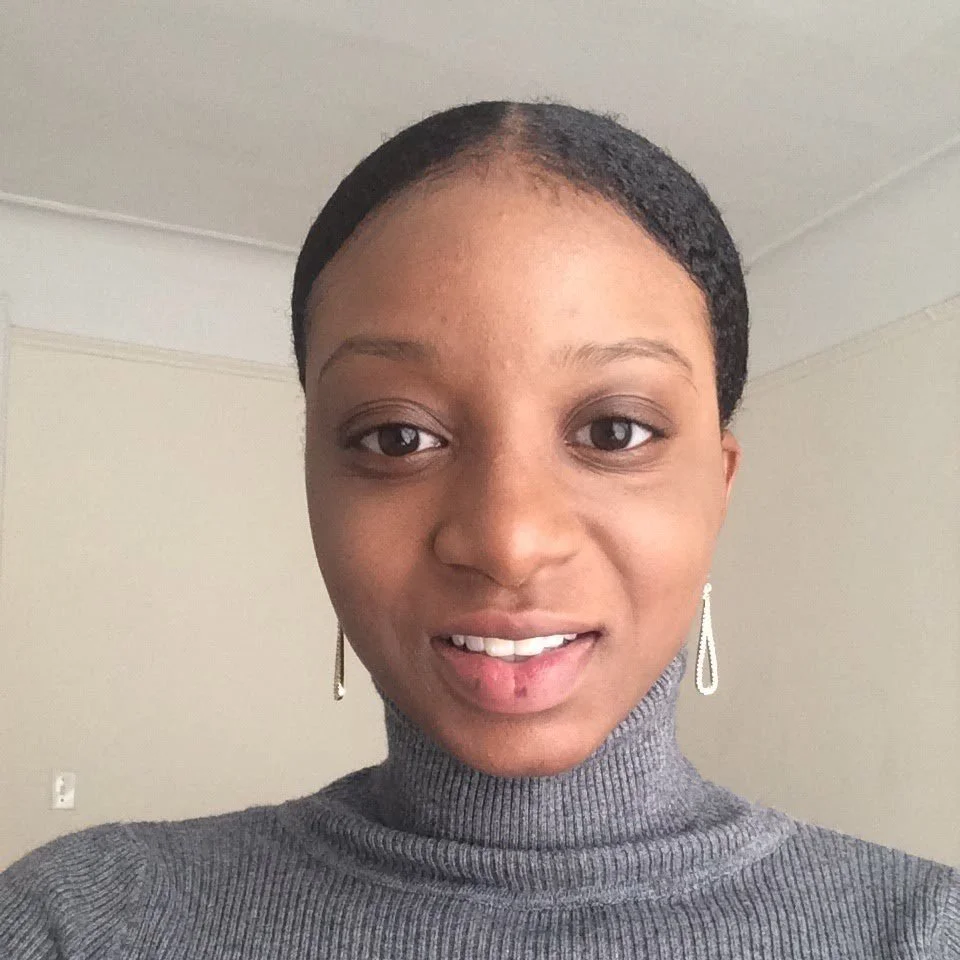 Close-up of a woman with short black hair, wearing silver earrings and a gray turtleneck sweater, smiling in a room with plain walls.
