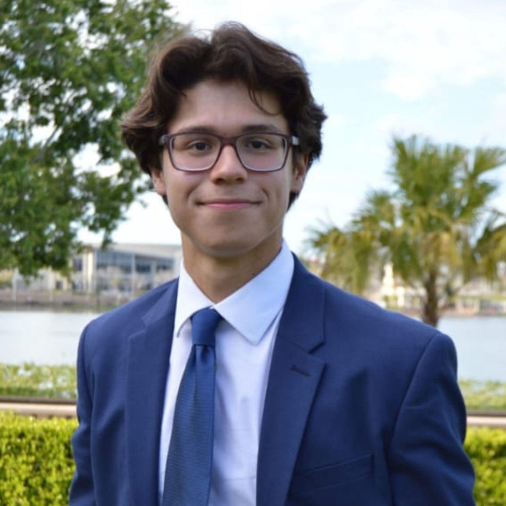Young man with brown hair, glasses, in a blue suit, white shirt, blue tie, outdoors near water, trees, and buildings, smiling at camera.