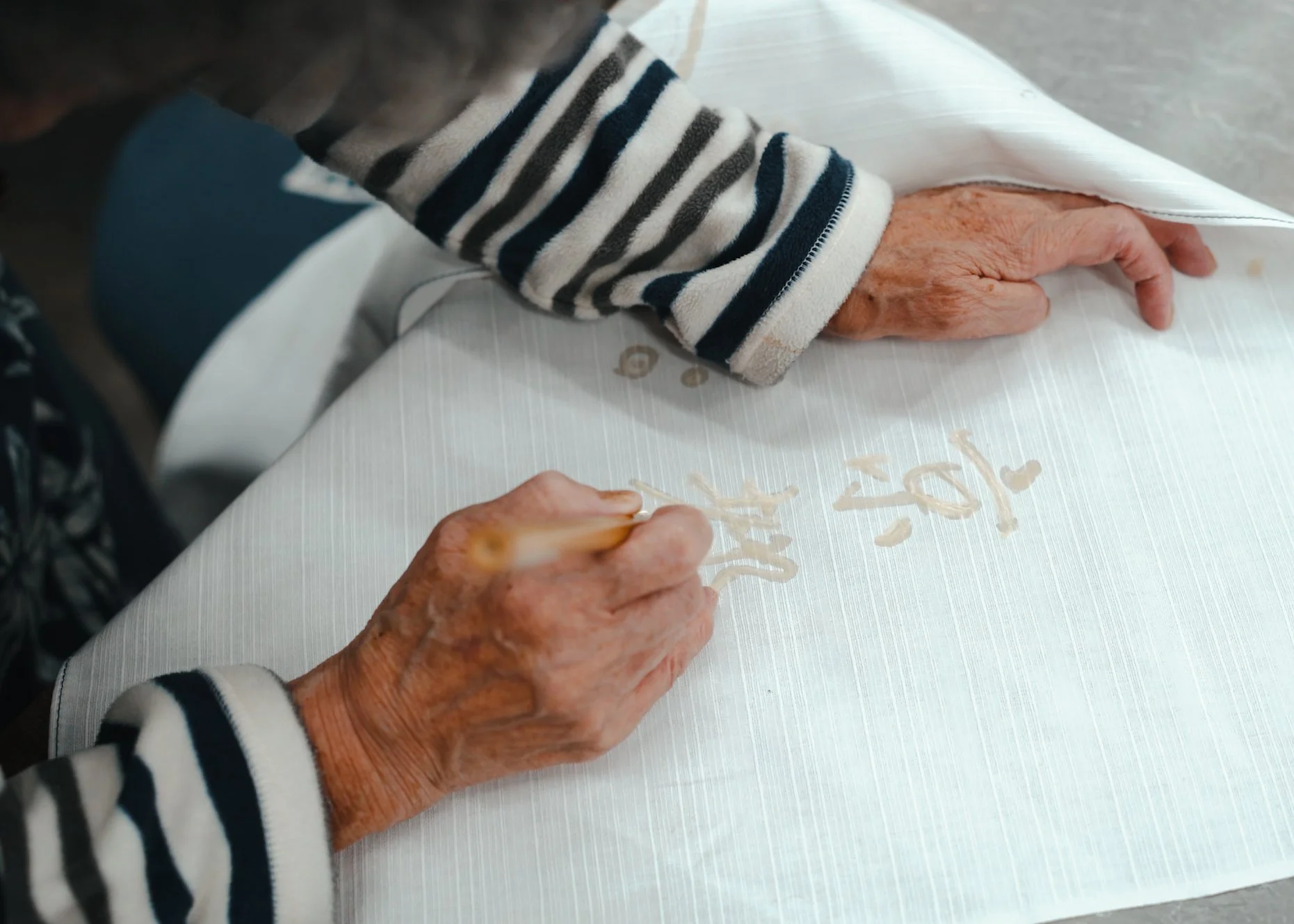 Elderly person with wrinkled hands writing on a white tablecloth with a gold-colored marker, wearing a striped long sleeve shirt.