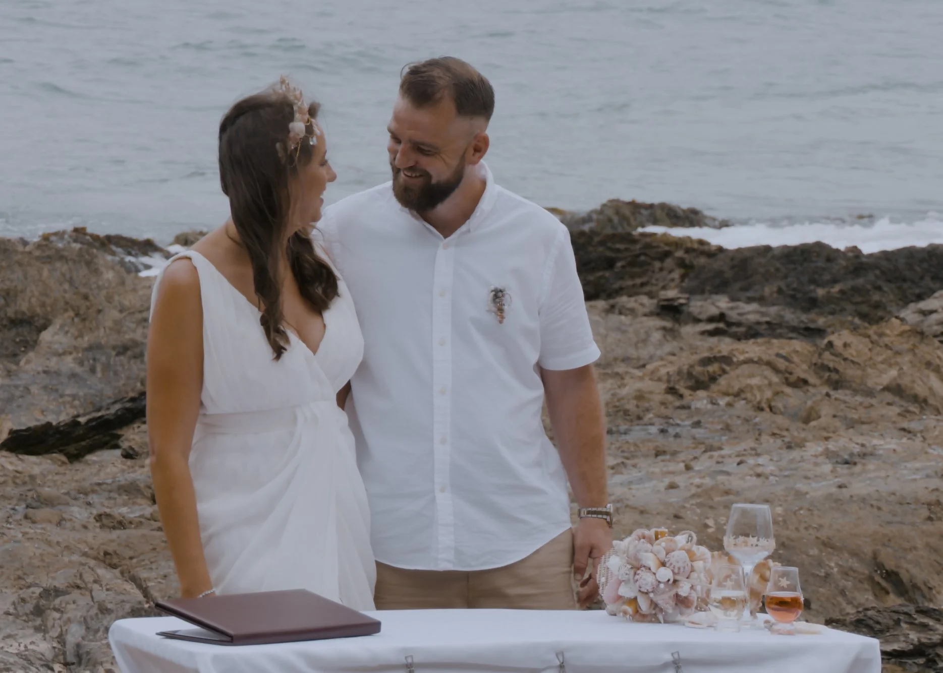 A couple standing on rocks by the ocean during their wedding, smiling at each other, with a table set with drinks and a shell bouquet.