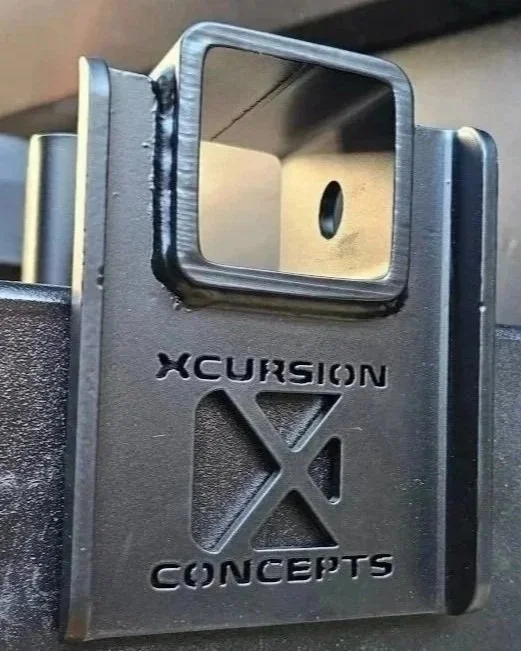 Close-up of a metal bracket or mount attached to a black surface, with the words 'XCursion X Concepts' embossed on it.