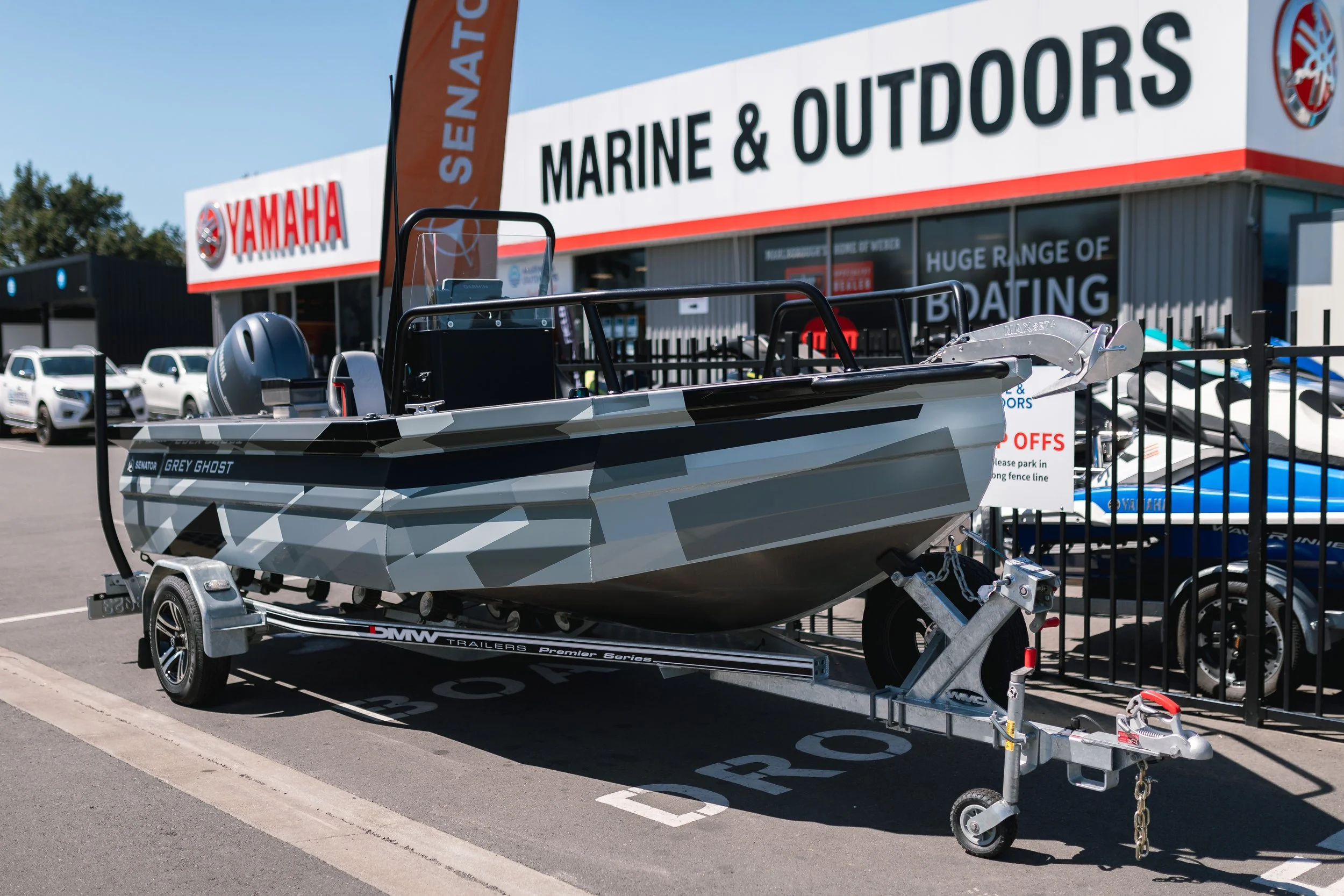 A grey camouflage-patterned boat on a trailer parked outside a marine and outdoors store. The boat has a small outboard motor at the stern and an aluminum propeller.