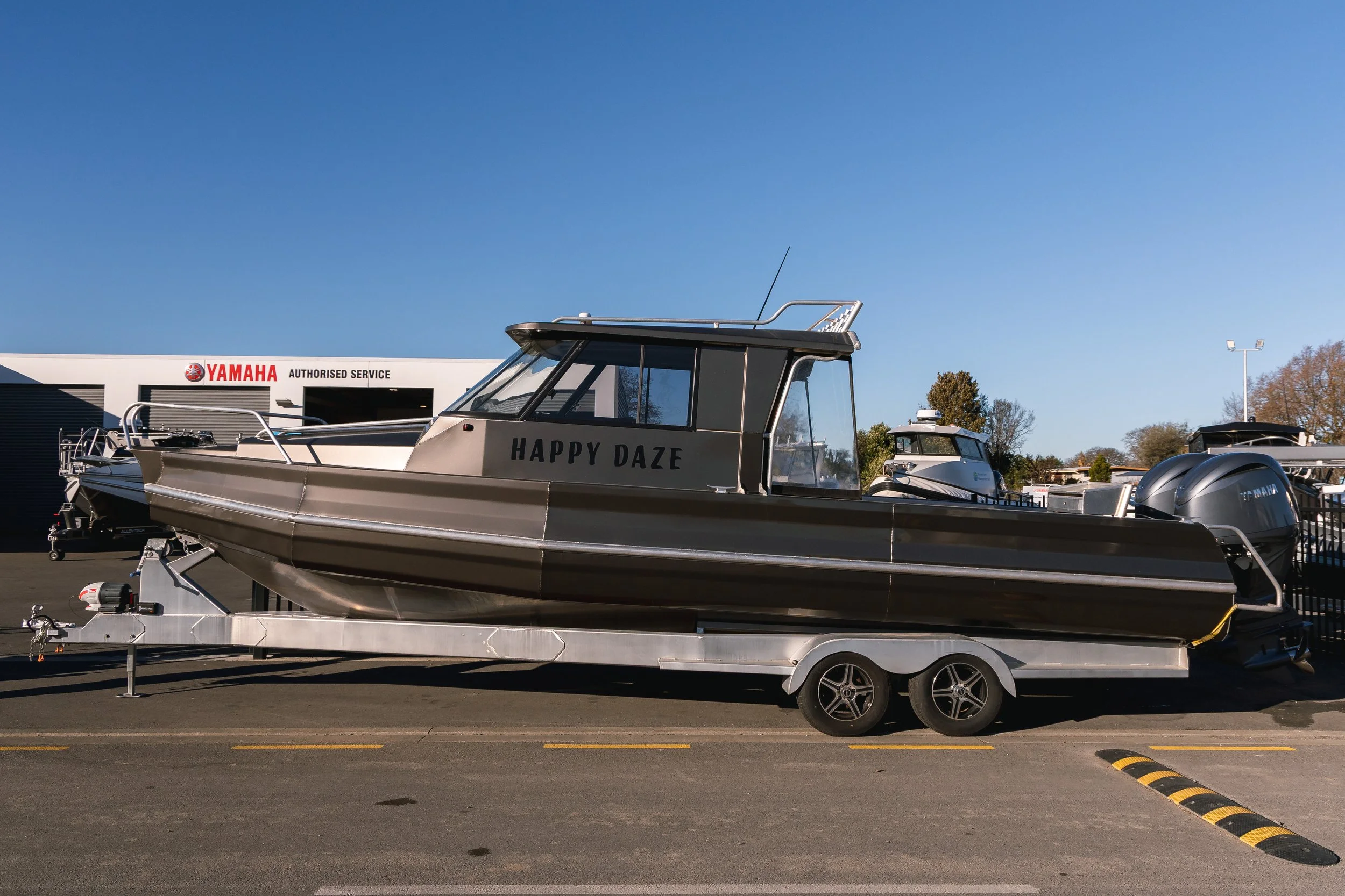 A boat named 'HAPPY DAZE' on a trailer in a parking lot, with a Yamaha authorized service building in the background and a clear blue sky above.