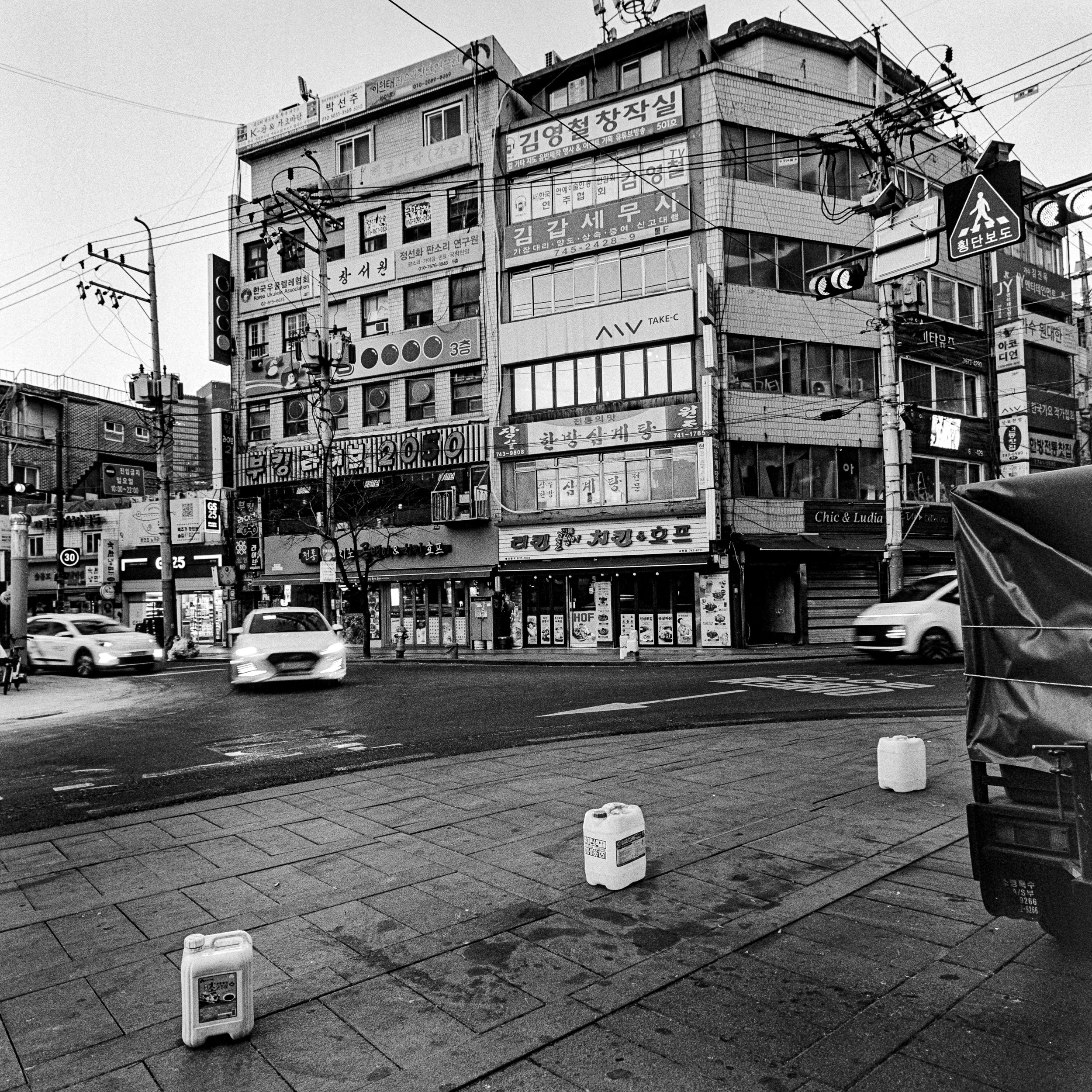Black and white photo of a busy street corner with moving cars, multi-story buildings with storefronts and signs in Korean, utility poles with wires, and traffic signals.
