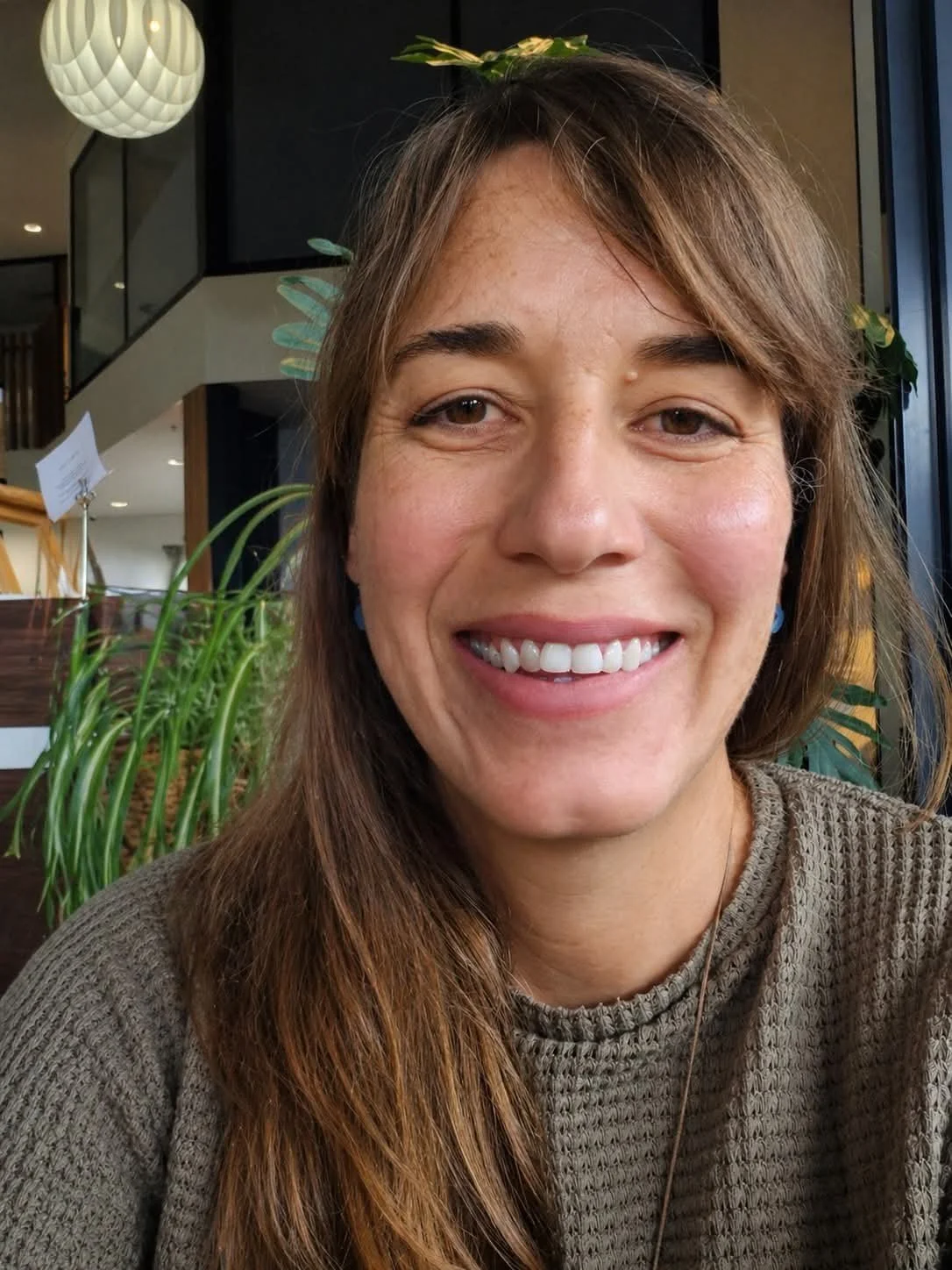A young woman smiling in a cafe with greenery and hanging lights in the background.