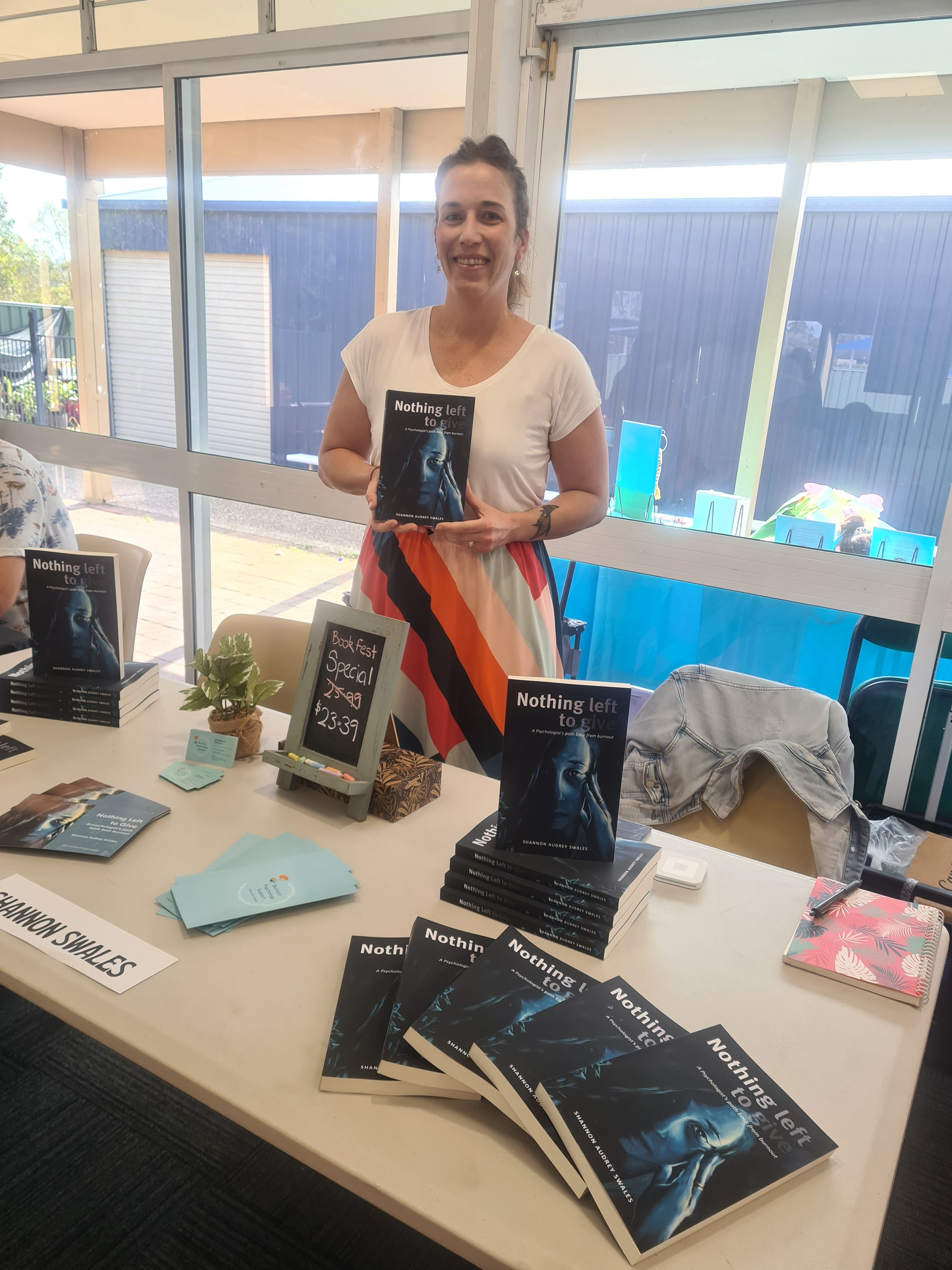 Shannon standing holding her book. In front of her is a table with her book and other merchandise. Book fair.