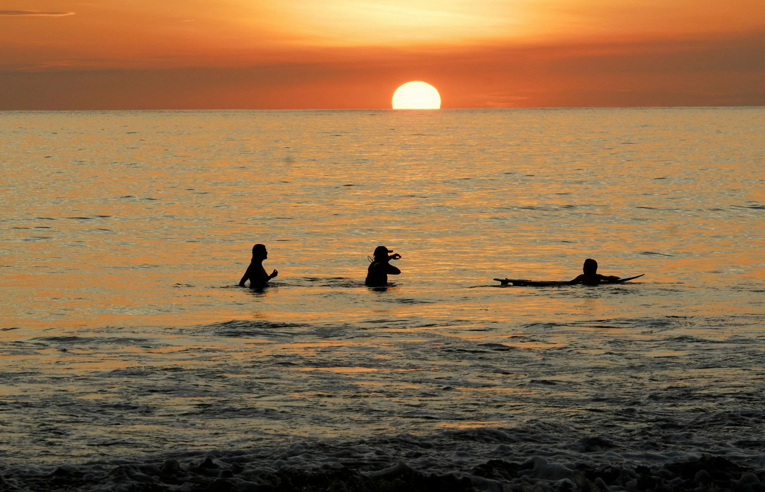 Three people enjoying sunset at the beach, one of them is paddleboarding while the other two are wading in the water, with the sun setting over the horizon.