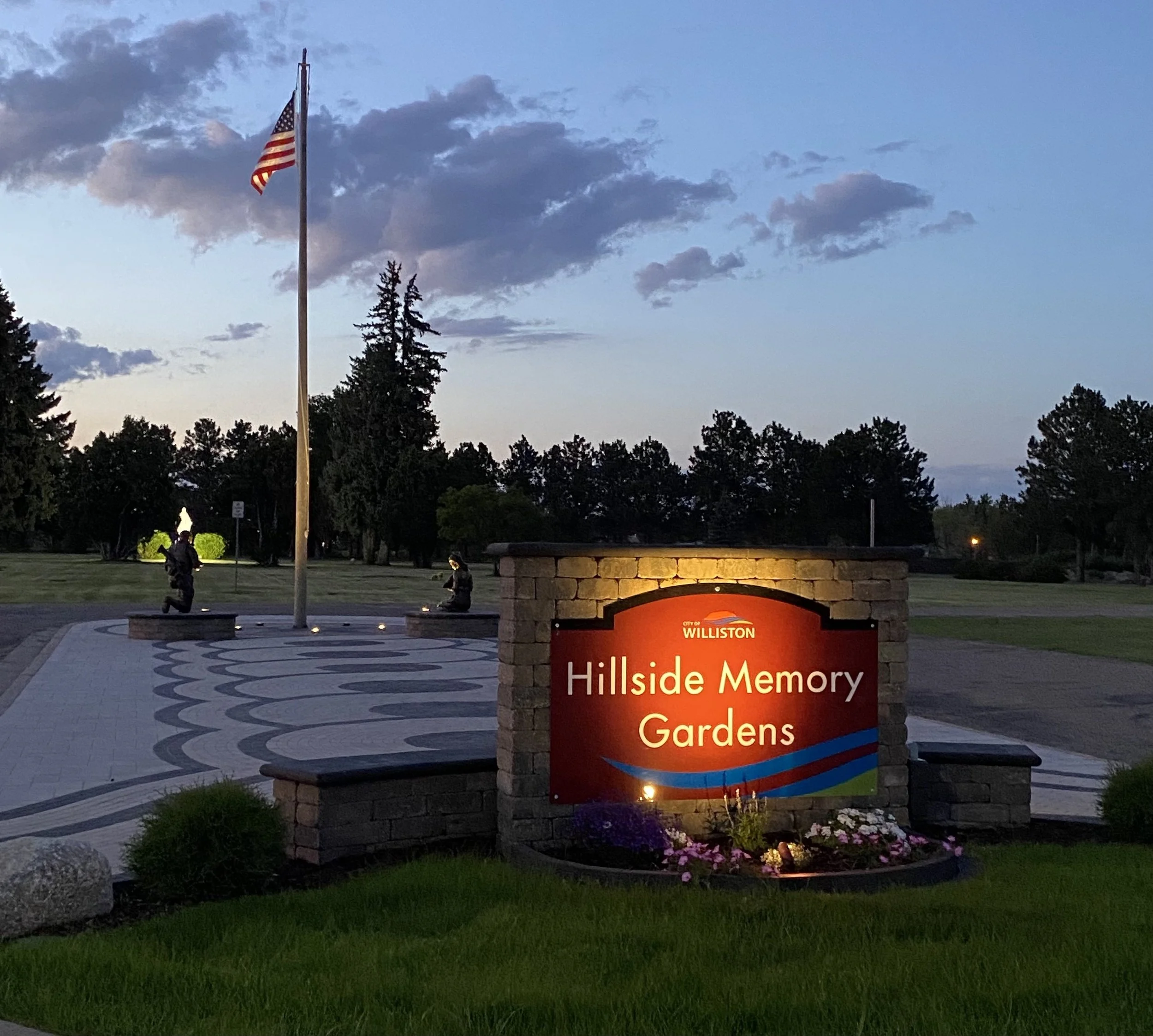 Entrance sign for Hillside Memory Gardens with an American flag on a tall flagpole, surrounded by framed landscaping, trees, and a clear blue sky.