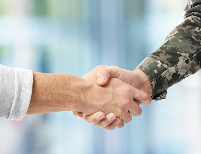 Two people shaking hands, one in a white shirt and the other in camouflage military uniform, in an indoor setting.