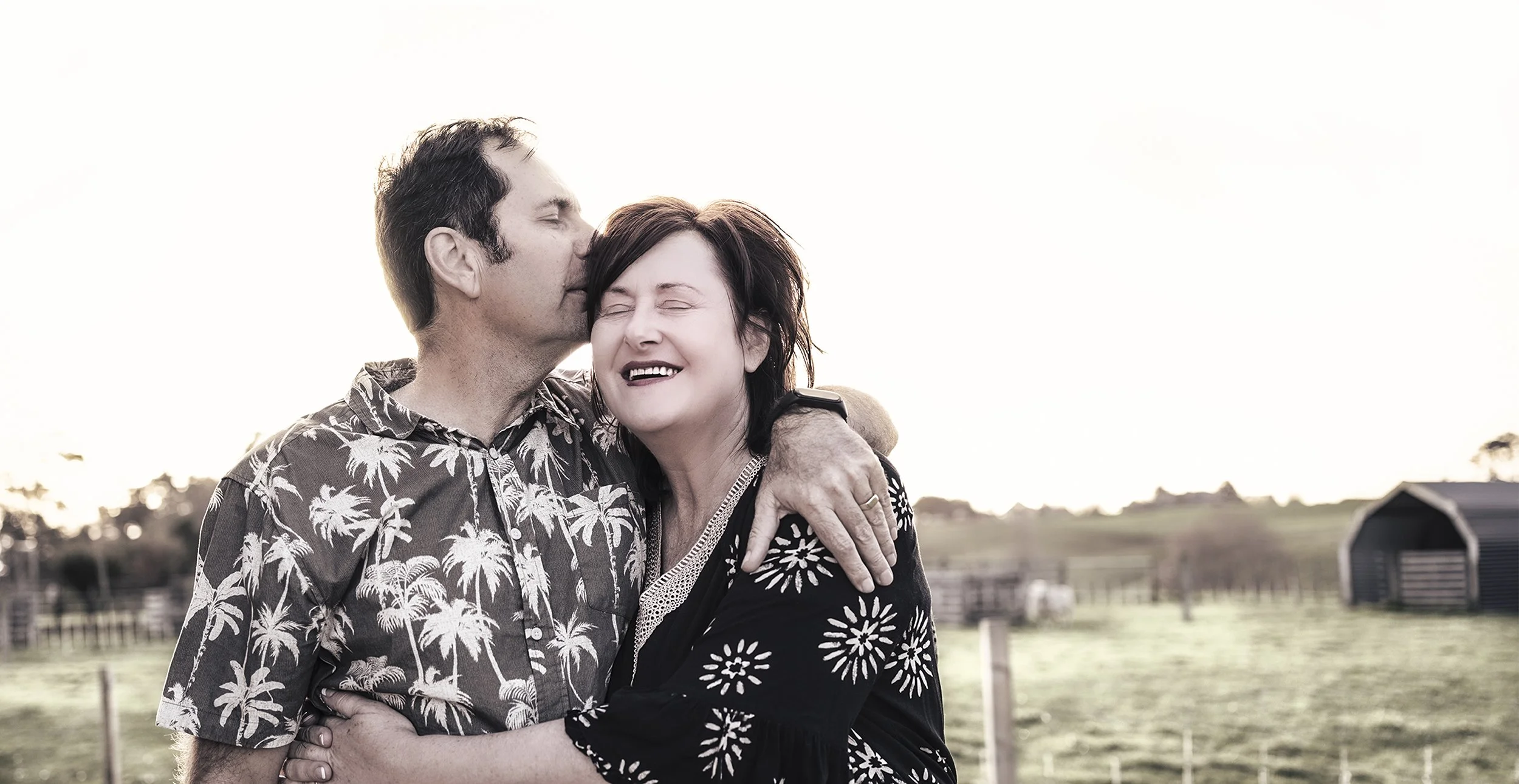 A man is kissing a woman on the forehead while they embrace outdoors in a rural area during sunset, with fields and a barn in the background.