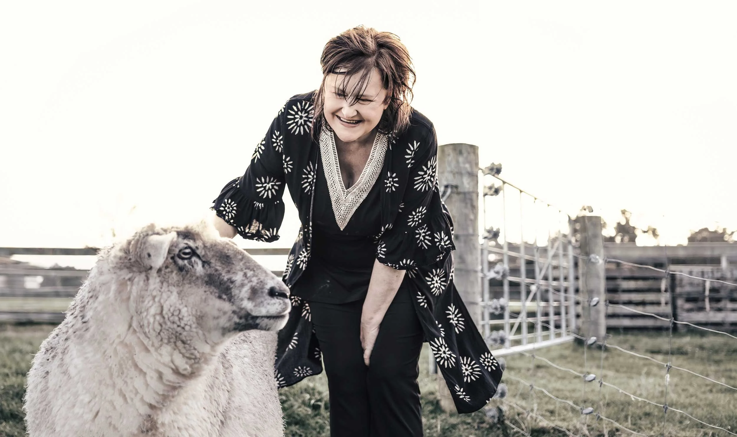 A woman smiling and leaning towards a sheep behind a fence outdoors.