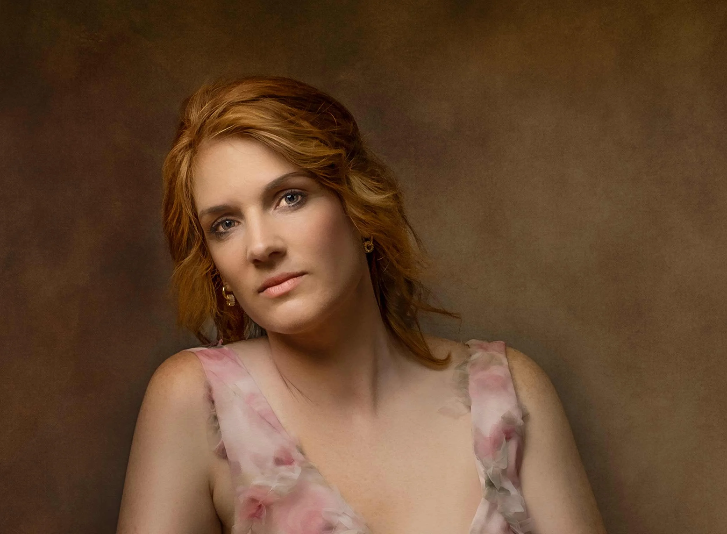Portrait of a woman with red hair, wearing a pink floral dress, looking at the camera against a brown background.