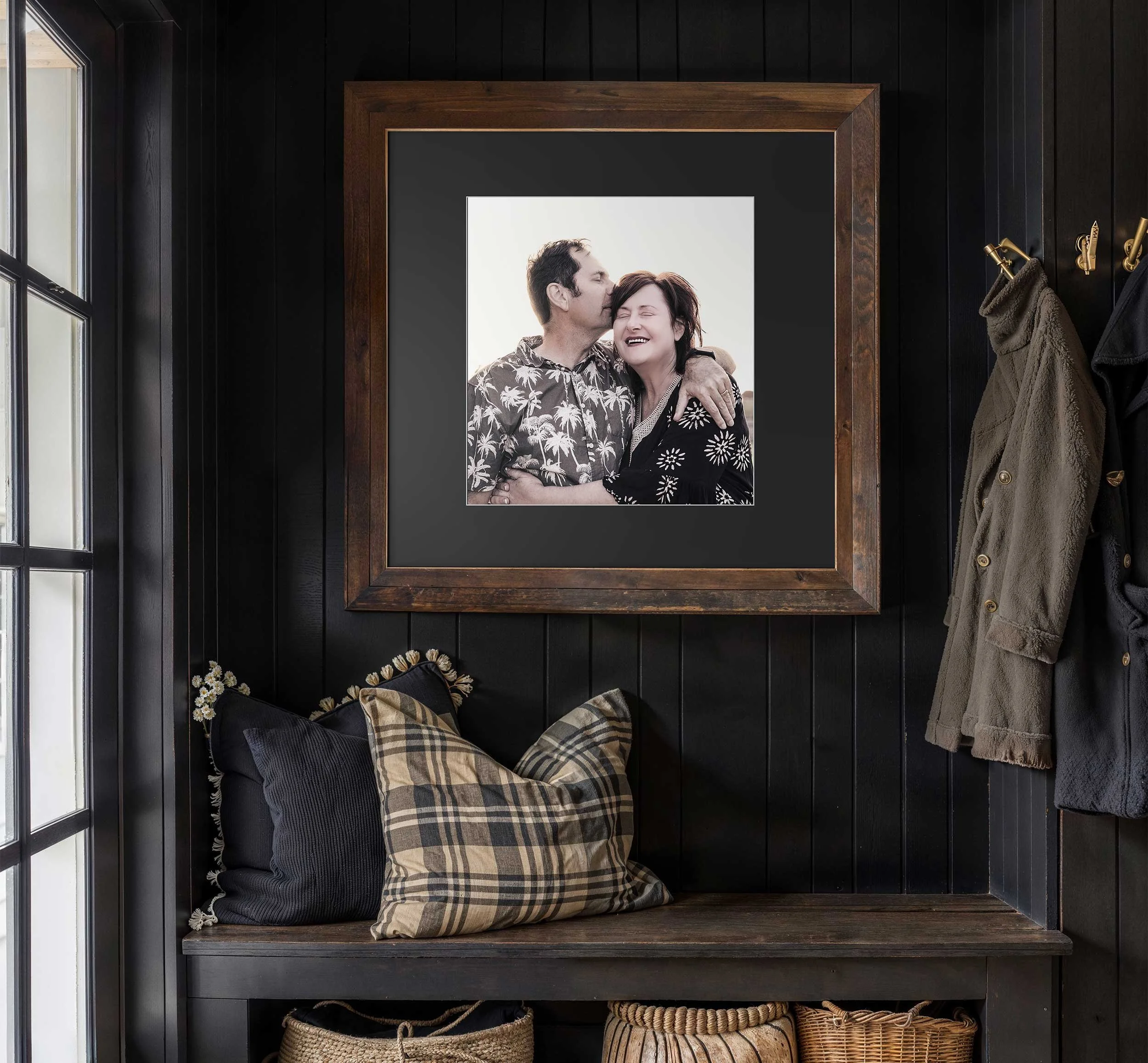 A framed black-and-white photograph of a smiling couple embracing, hanging on a dark wooden wall in a cozy entryway with a bench, pillows, and hanging coats.