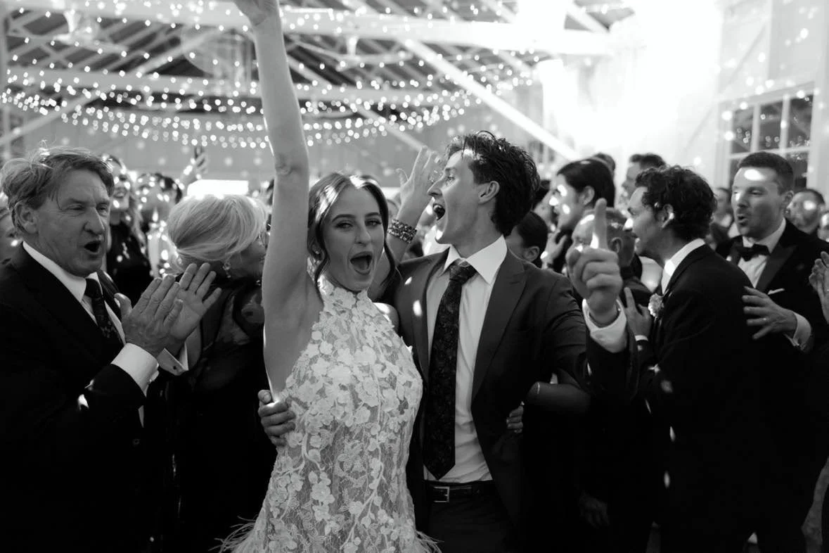 People celebrating at a wedding reception. A bride and groom are dancing and singing, surrounded by guests clapping and enjoying the party.