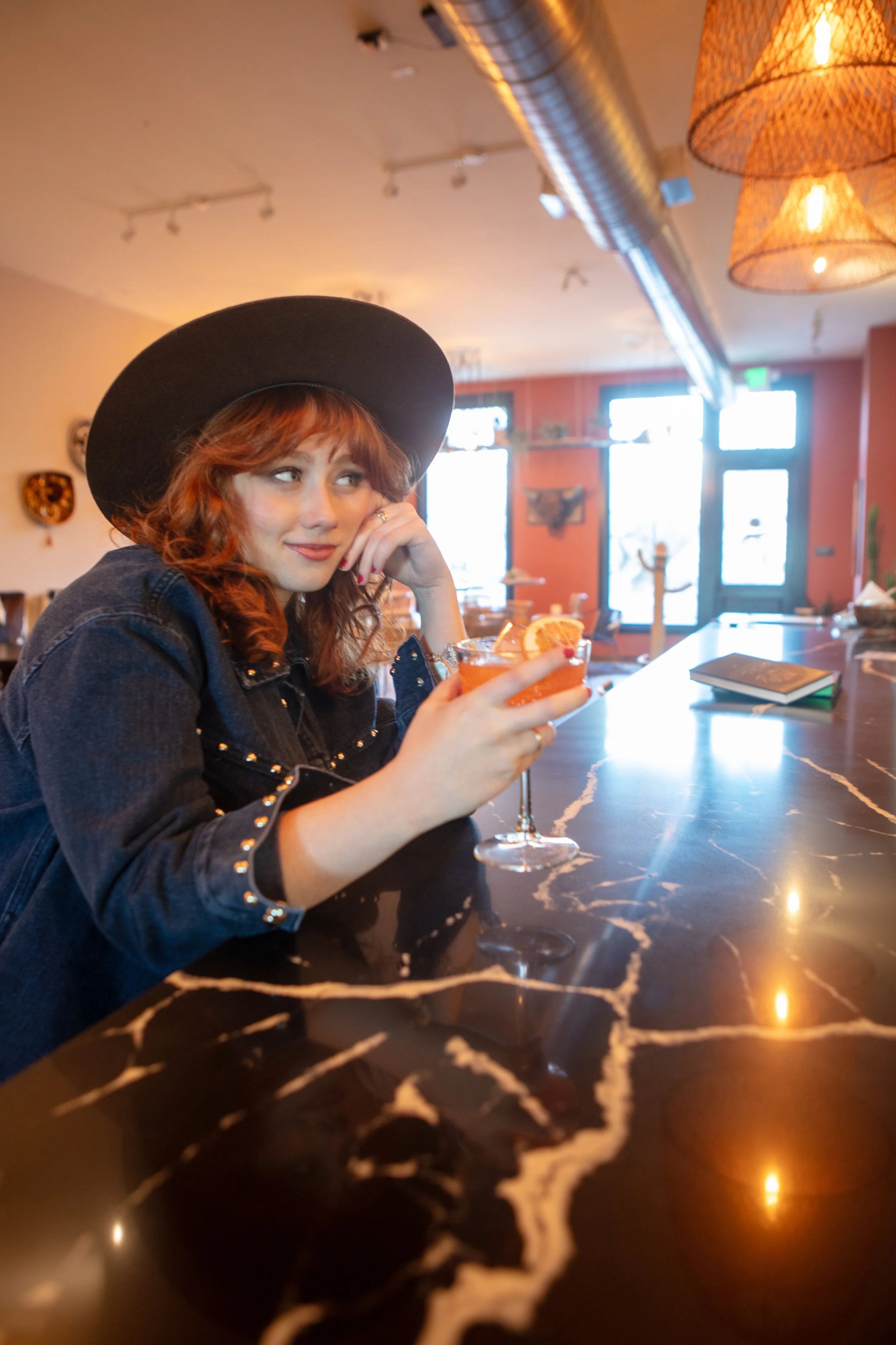 A woman with red hair wearing a black hat and denim jacket sitting at a marble bar counter, holding a cocktail with orange slice garnish, in a warmly lit restaurant or bar.