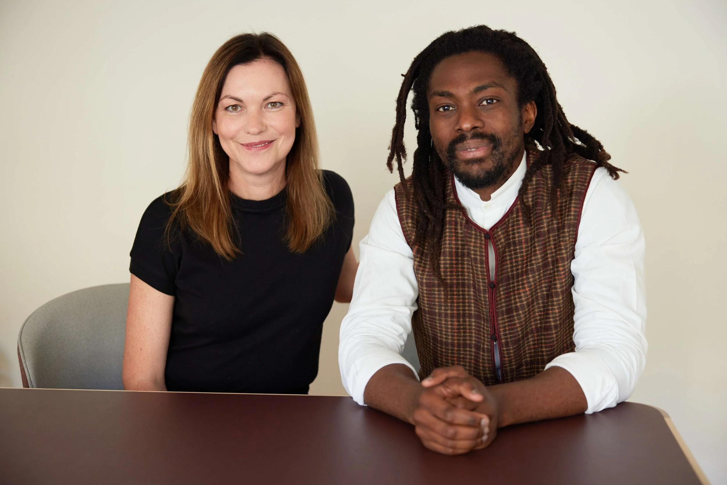 A woman with long brown hair and a black top sitting next to a man with dreadlocks, a beard, and a white shirt with a brown checkered vest, both smiling and sitting at a brown table in front of a plain light-colored wall.