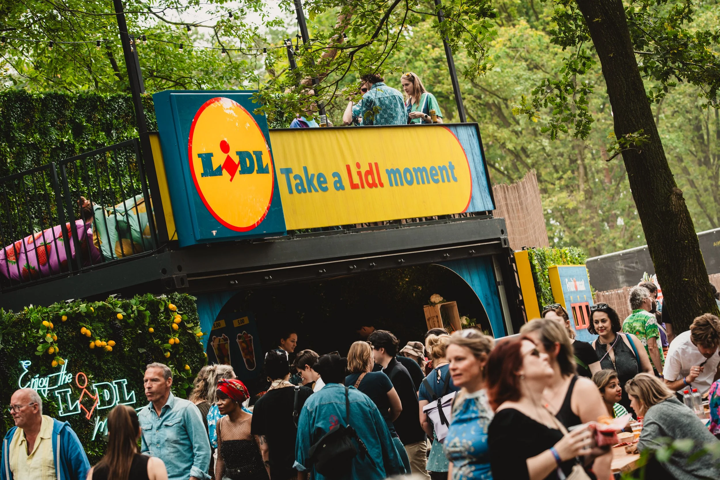 Drukke markt met mensen onder bomen, een zonnige dag, een stand met een Lidl-banner die zegt: 'Take a Lidl moment'.