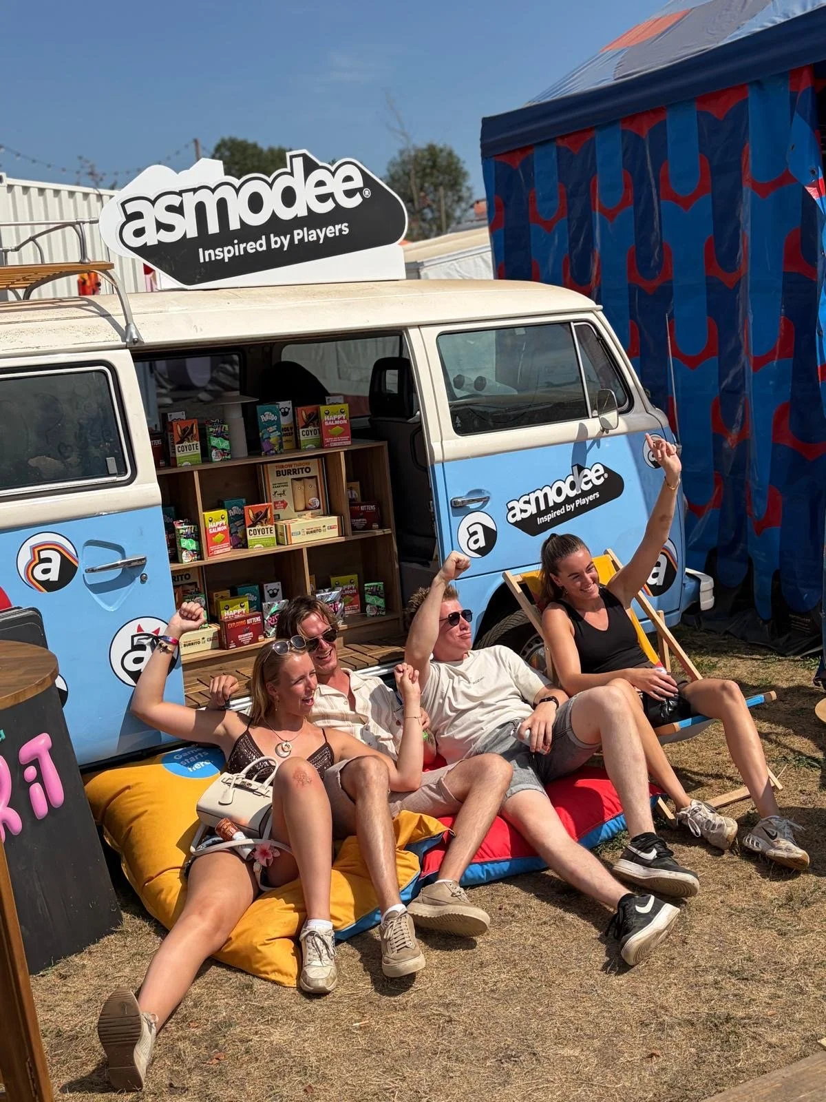 Vijf mensen die ontspannen op een matje zitten, voor een blauwe foodtruck met het logo 'asmodee' en boekenrek, in een festivalsetting met een blauwe lucht en een grote blauwe en rode tent op de achtergrond.