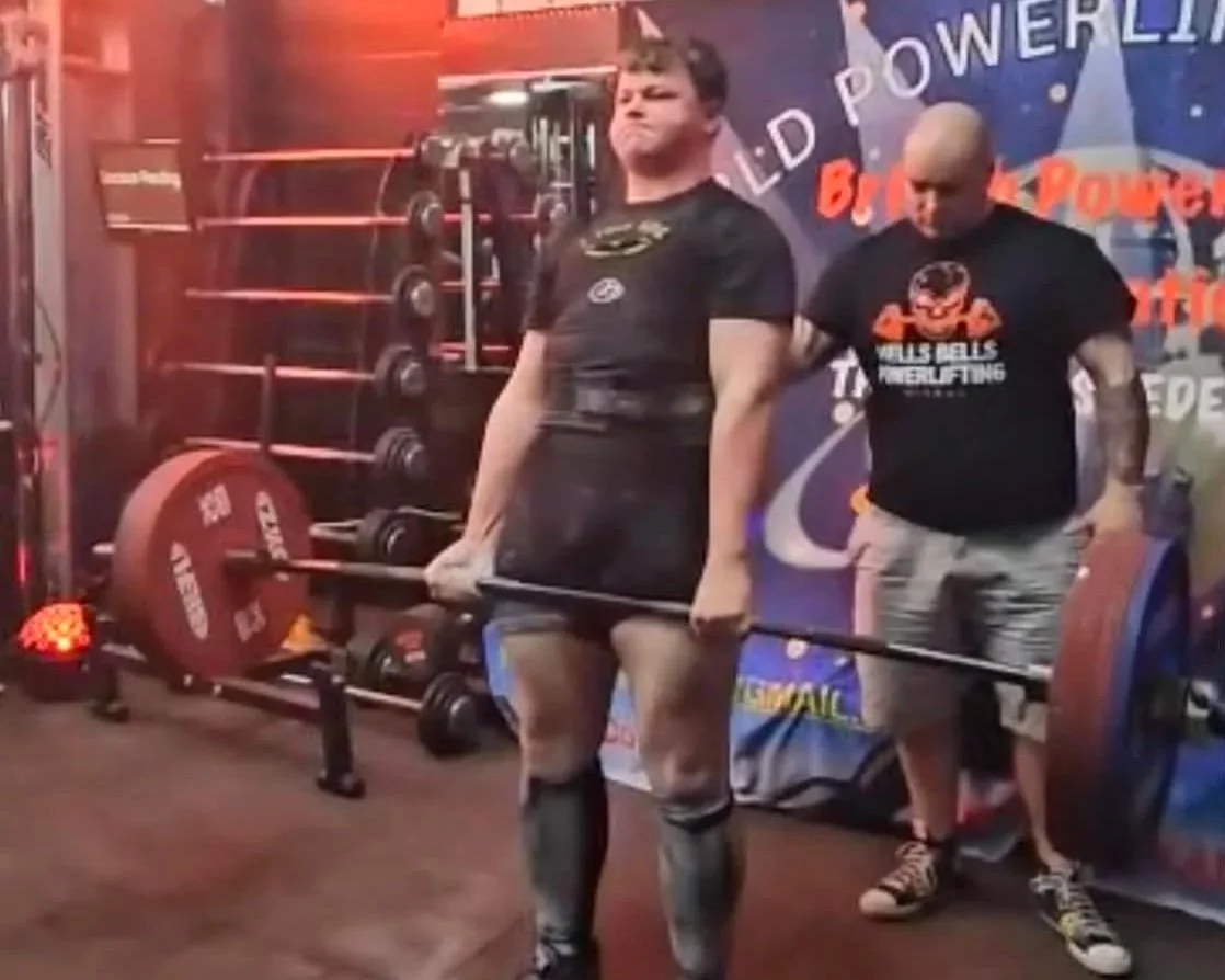 A woman lifting a heavy barbell with weights at a powerlifting competition, with a man standing behind her in a sleeve-less shirt, and a background featuring powerlifting banners and equipment.