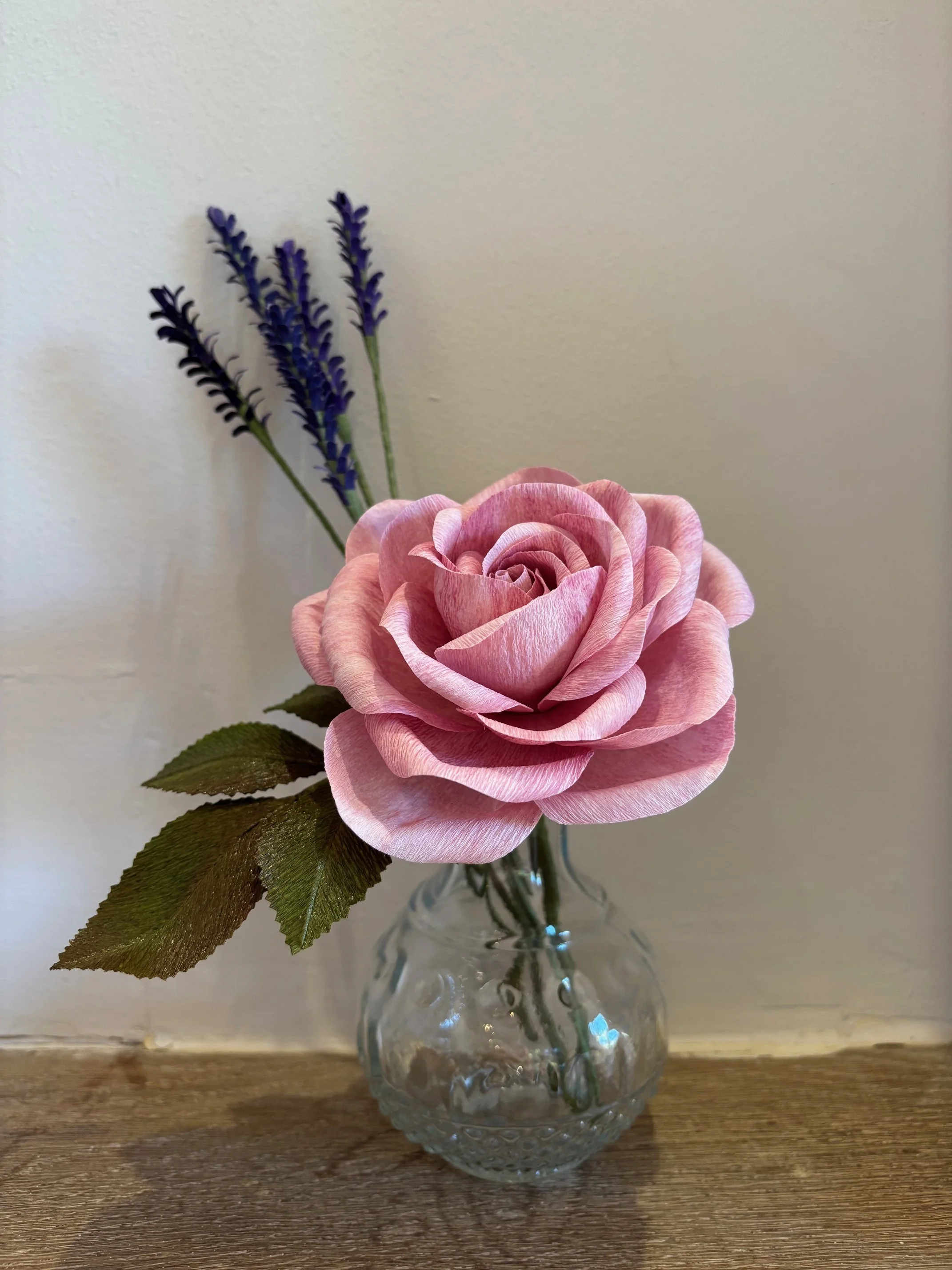 Pink paper rose with green leaves and purple paper lavender stems in a textured glass vase on a wooden surface against a plain wall.