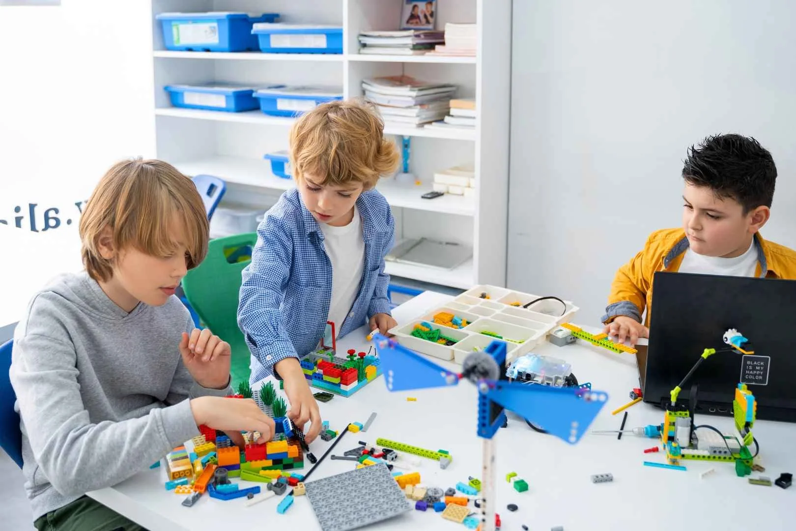 Drie kinderen werken samen aan een bouwproject met Lego-stenen op een tafel, er is een computer en bouwmateriaal aanwezig.