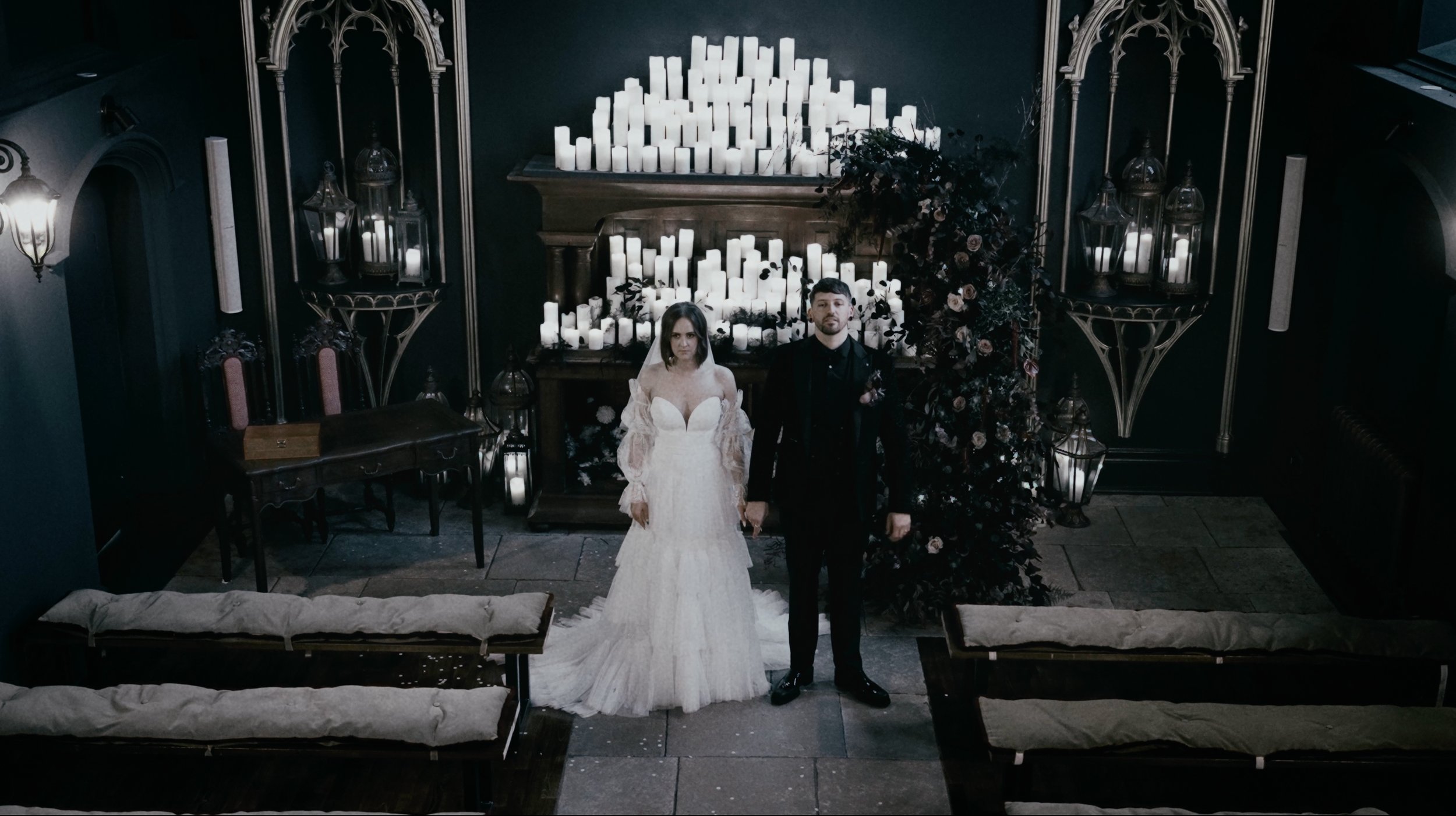 A couple stands in a dimly lit room, dressed in wedding attire. The bride is wearing a white gown with lace sleeves, and the groom is in a dark suit. They hold hands in front of an altar adorned with numerous lit candles. The setting has a gothic sty