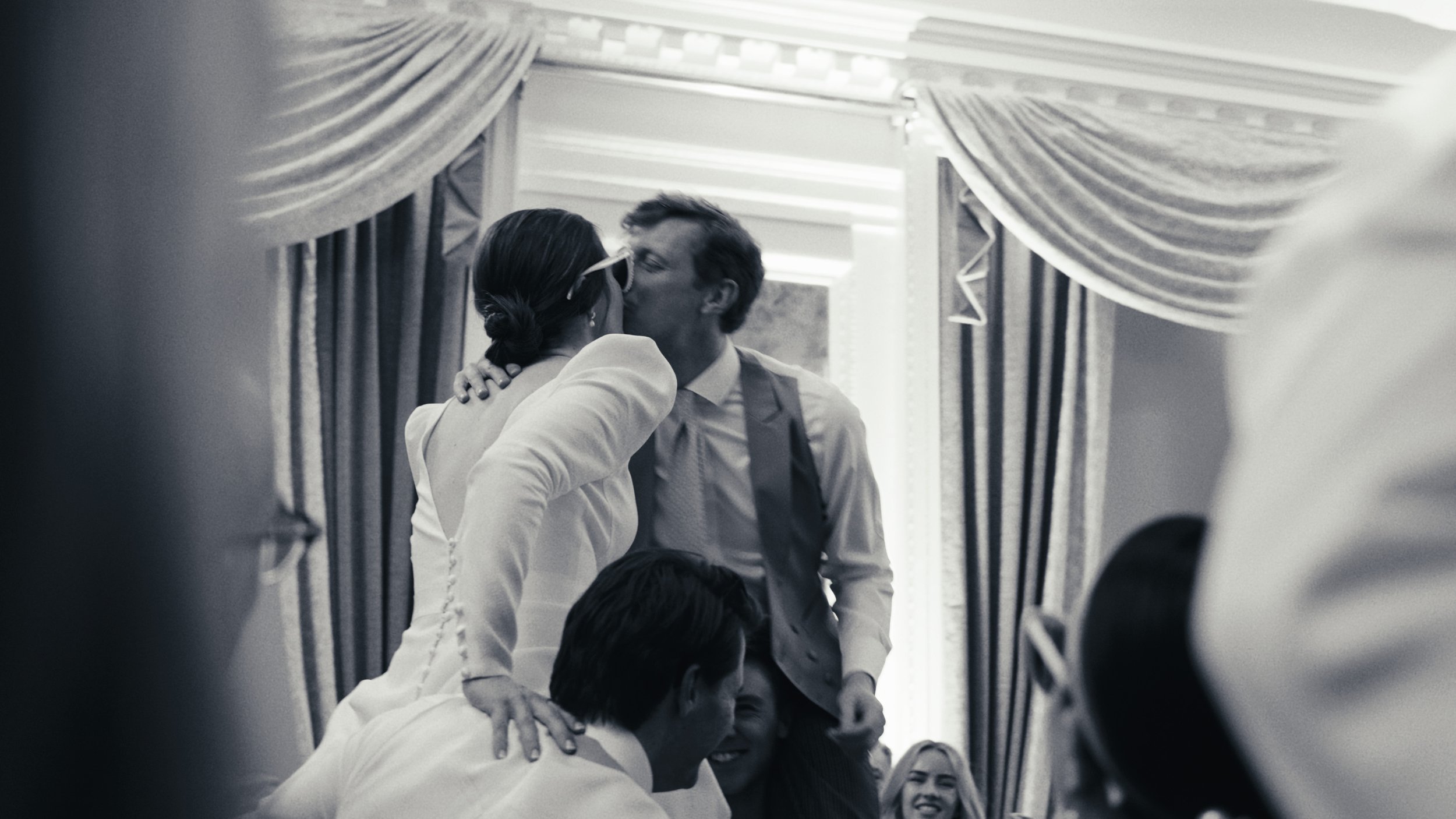 Black and white photo of a couple kissing at a wedding, surrounded by people. The woman is wearing a dress with buttons and sunglasses. The man is in a vest and tie, leaning in for the kiss. Curtains and ornate wall detailing are in the background.