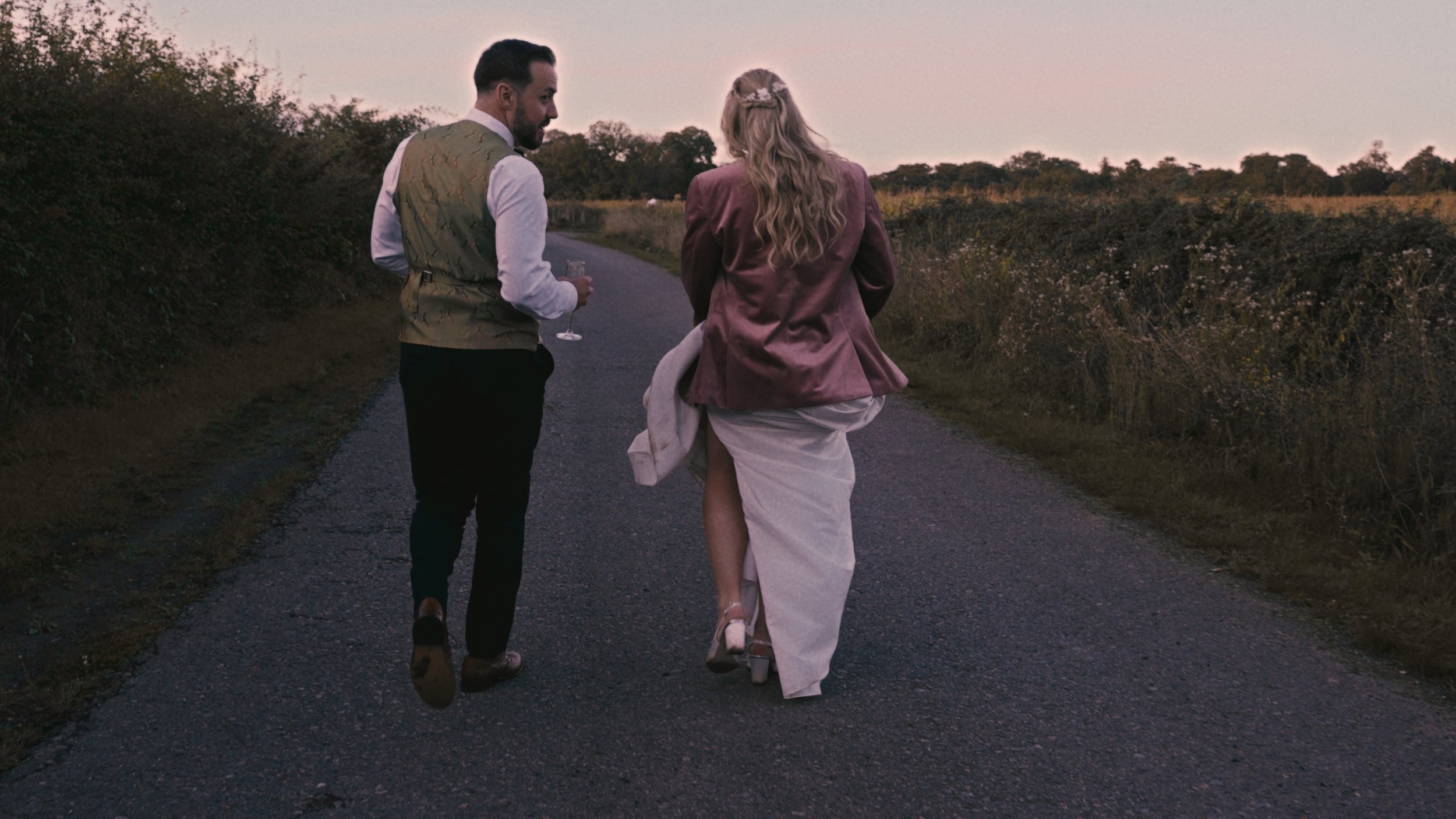 Bride and groom walking at sunset whilst bride wears groom's jacket.