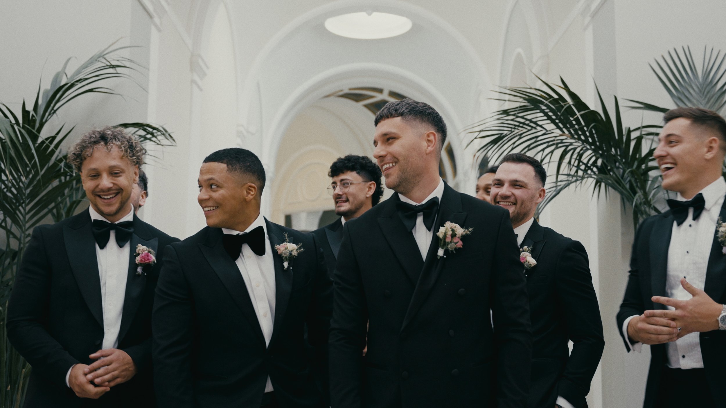 Group of groomsmen in black tuxedos with bow ties and boutonnieres, smiling and standing indoors with palm plants in the background.