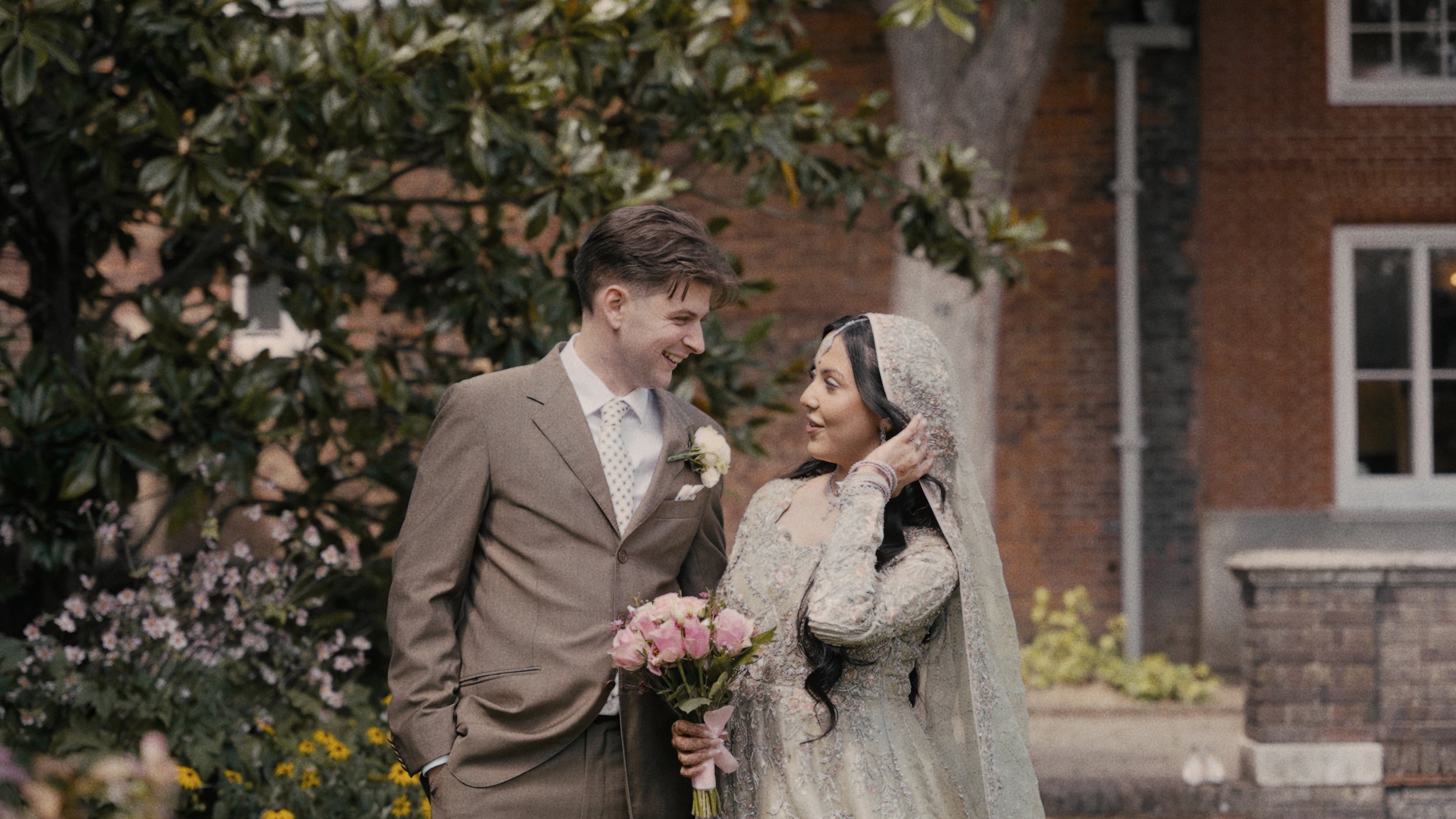 Bride and groom standing outdoors in garden, smiling at each other. Background features greenery and a brick building.