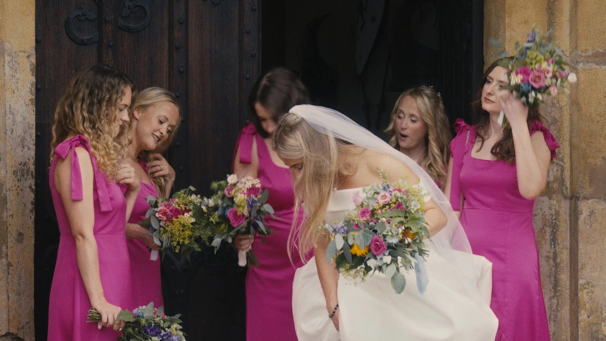 Bride and bridesmaids in pink dresses holding bouquets, standing outside a door, smiling and interacting.