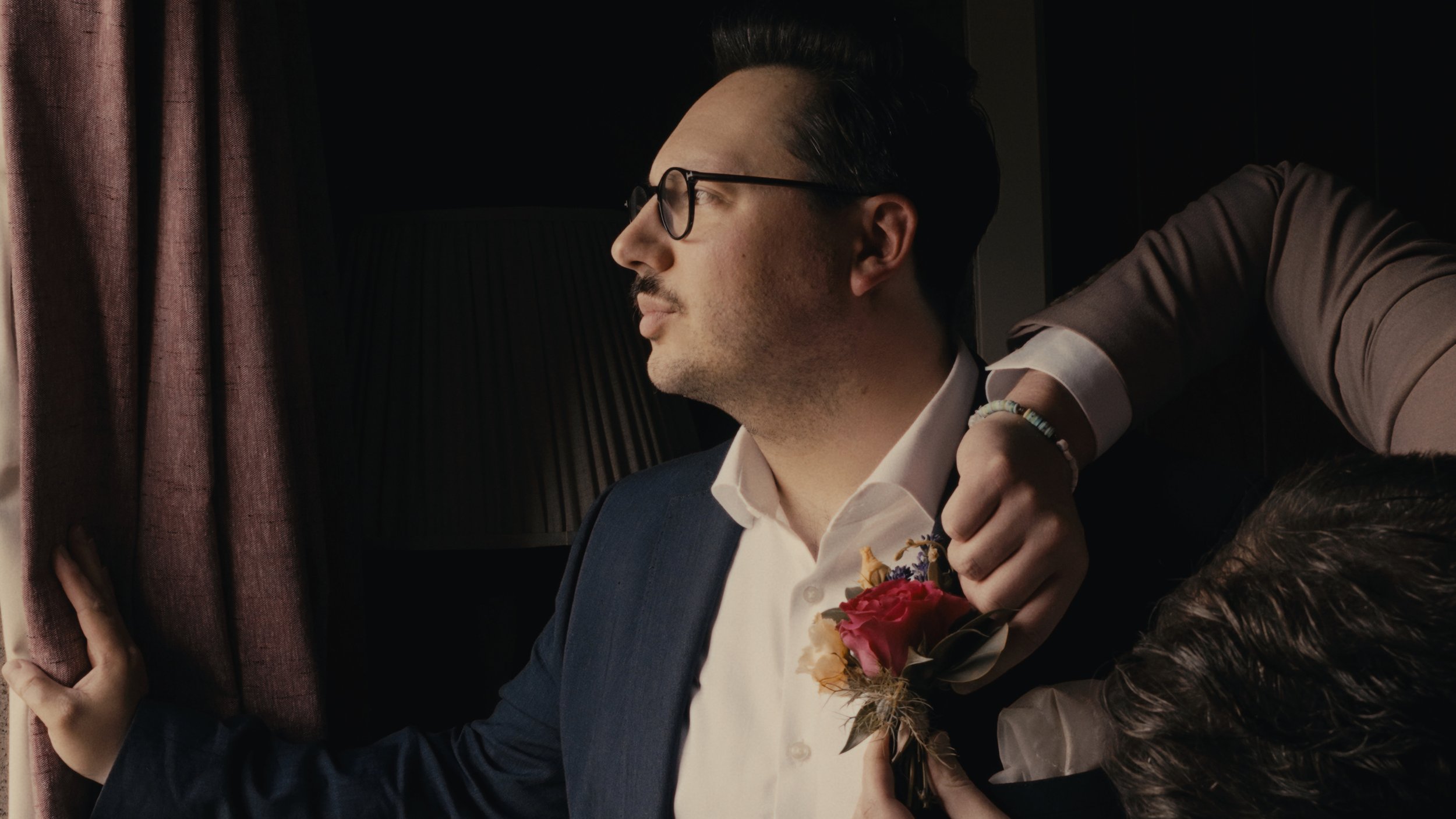 Groom standing at a window getting his button hole put on