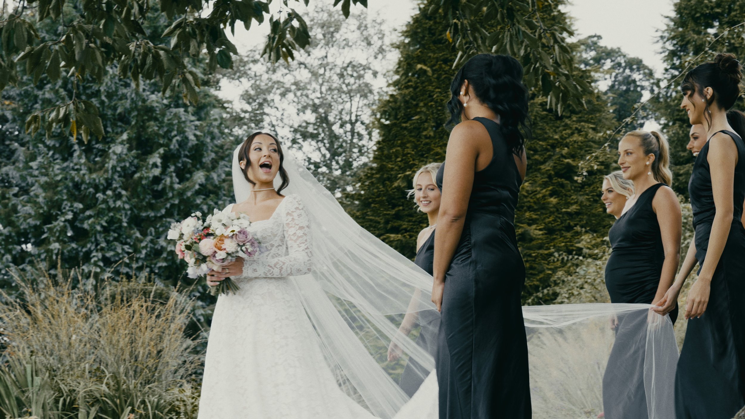 Bride in white dress surrounded by bridesmaids in black dresses, holding bouquet, outdoors with trees in background.