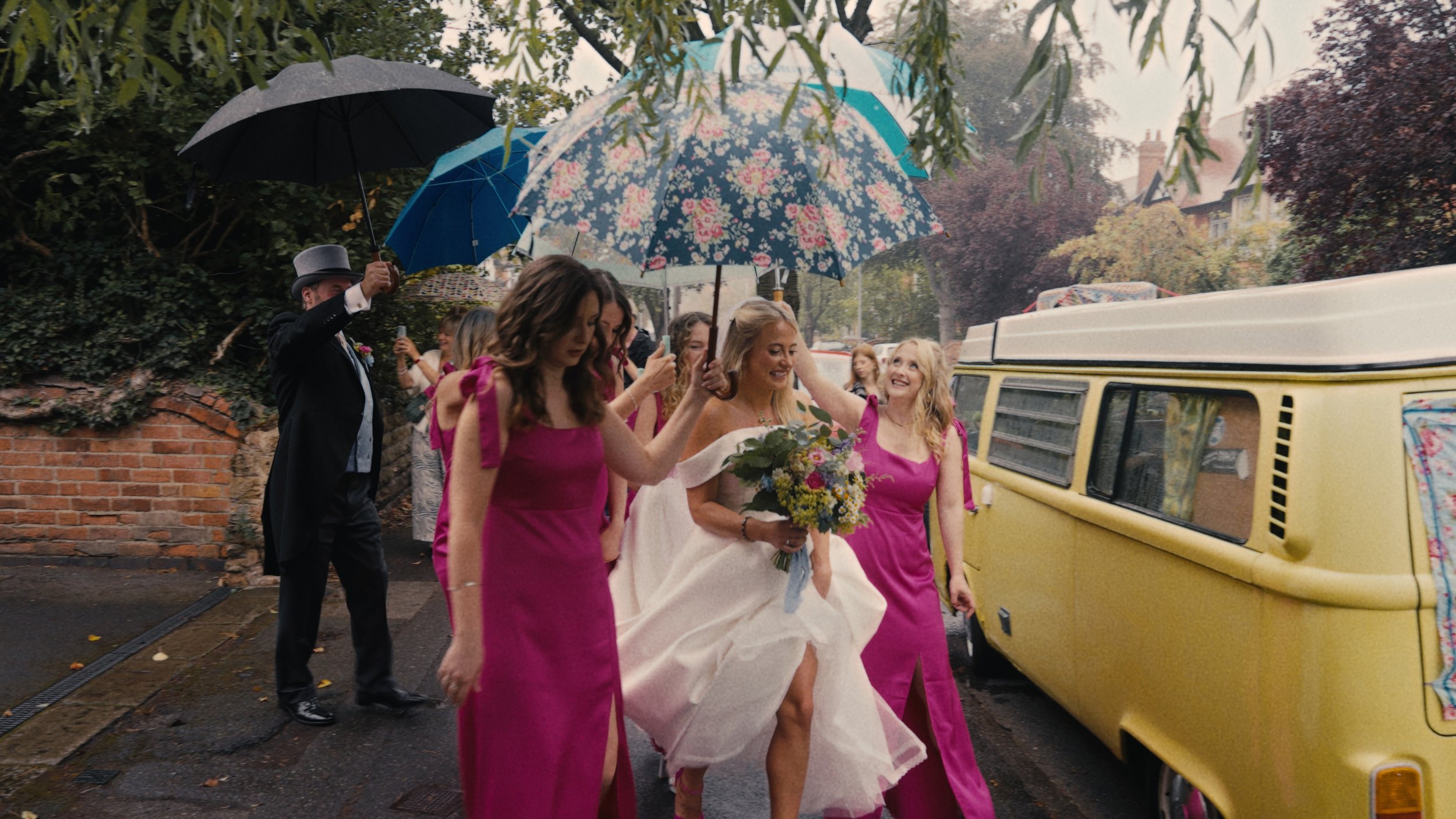 Bridesmaids in bright pink dresses holding umbrellas over the bride walking to a camper van