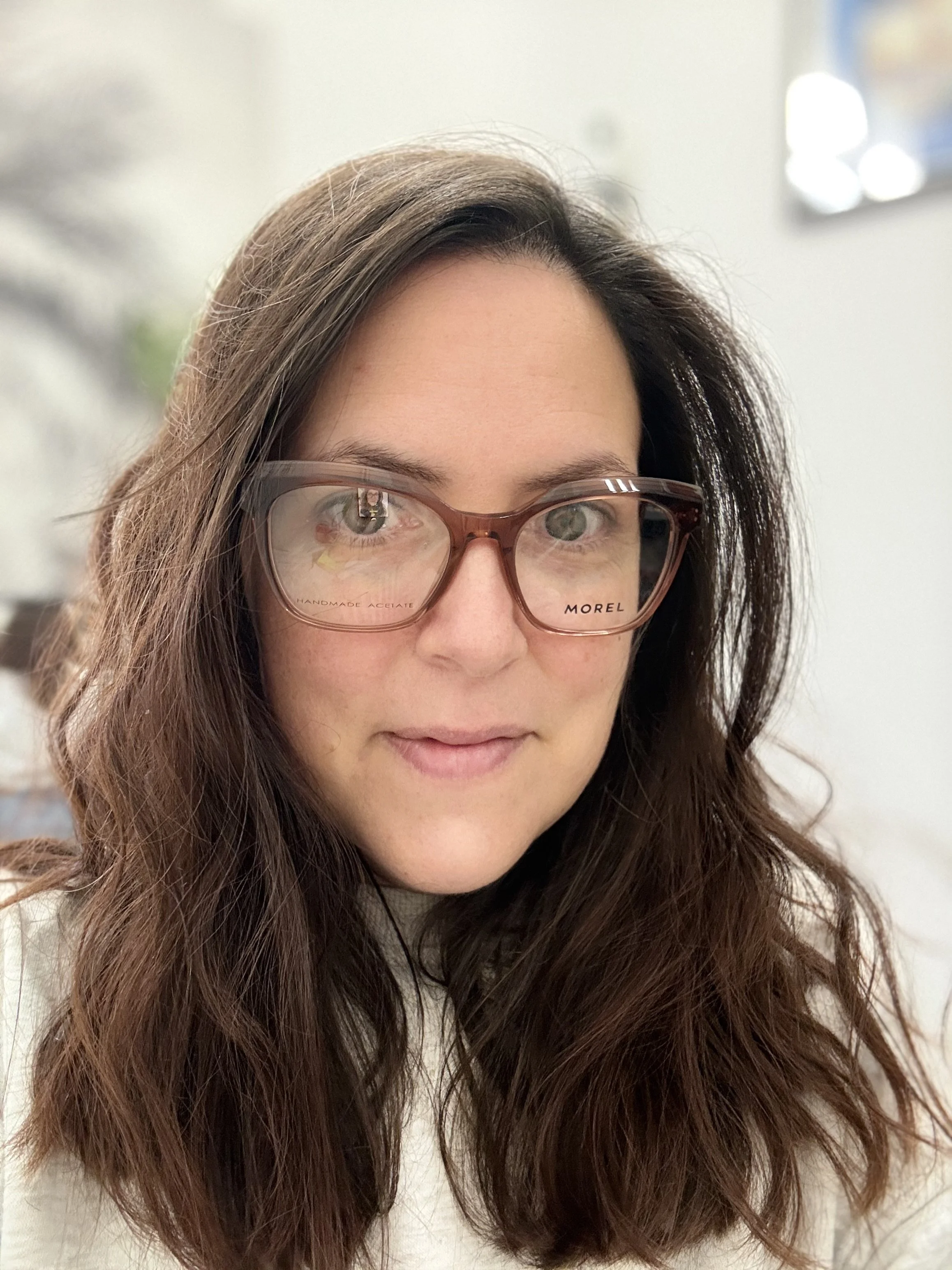 Close-up of a woman with long brown hair and glasses, smiling softly, inside a bright room. Morel szemüvegkeret a Showroom Optikában Budapesten. Barna Morel női keret.