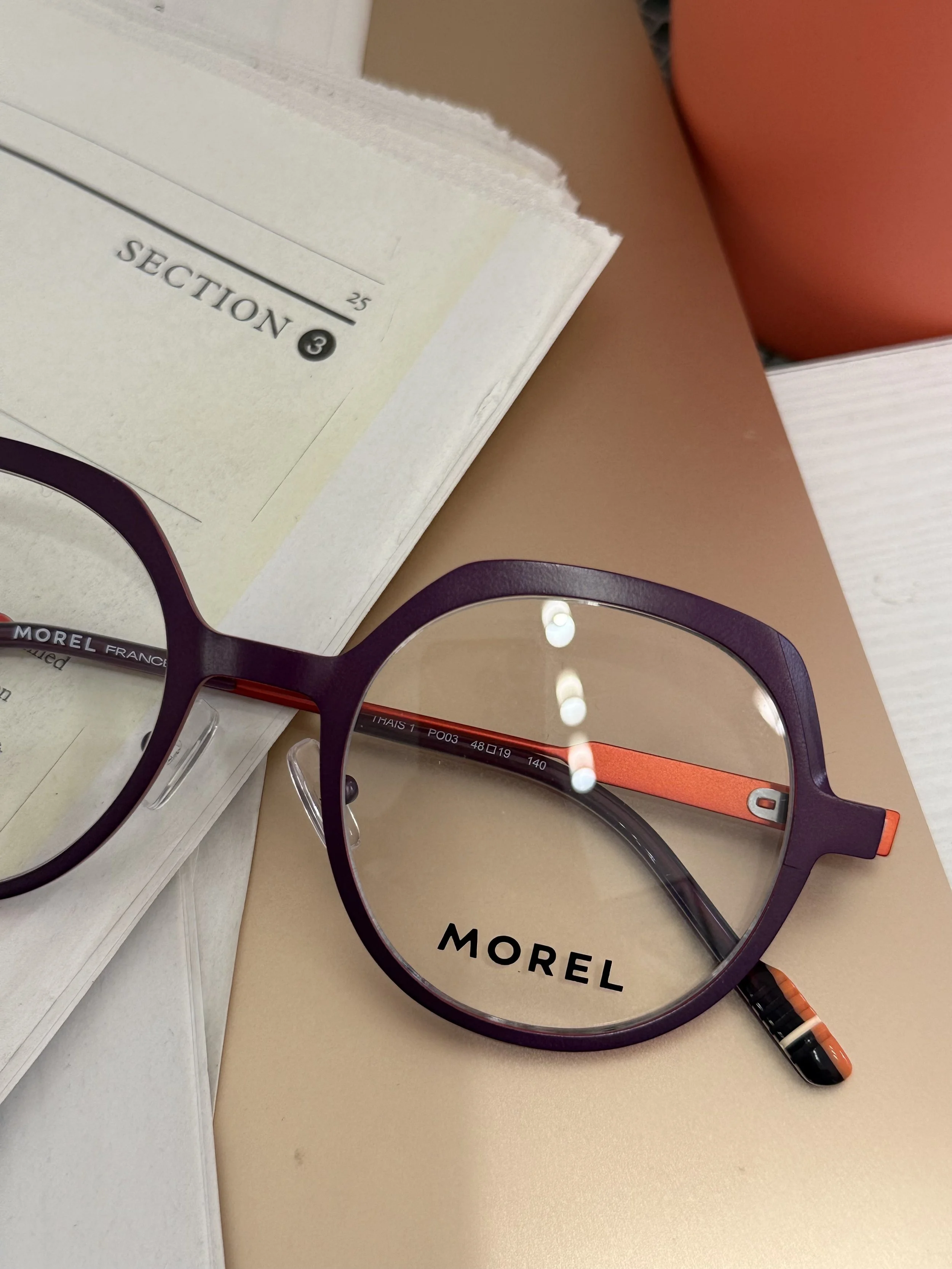 A pair of black and orange glasses resting on a beige surface near a white book with the text 'SECTION 8' on the page. Morel szemüvegkeret a Showroom Optikában Budapesten.